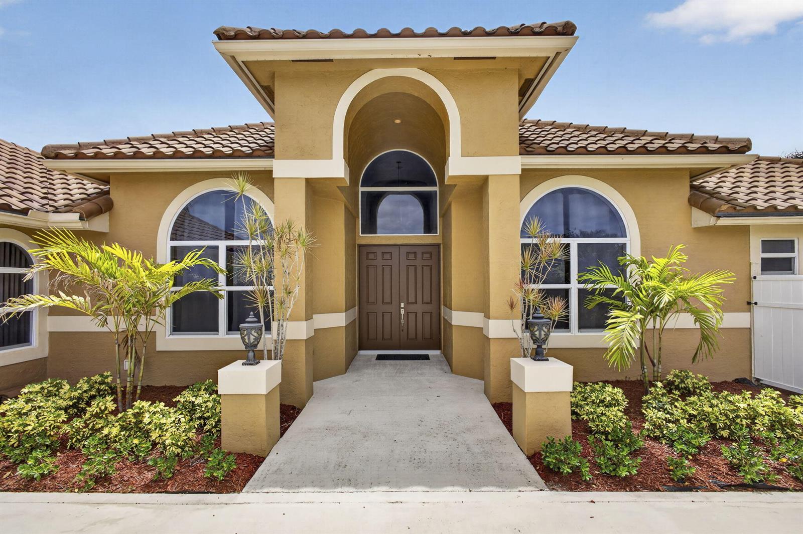 The exterior of a tan stucco house features a double front door and arched windows — 8030 Woodsmuir Drive, Palm Beach Gardens FL 33412, PALM BEACH GARDENS, FL
