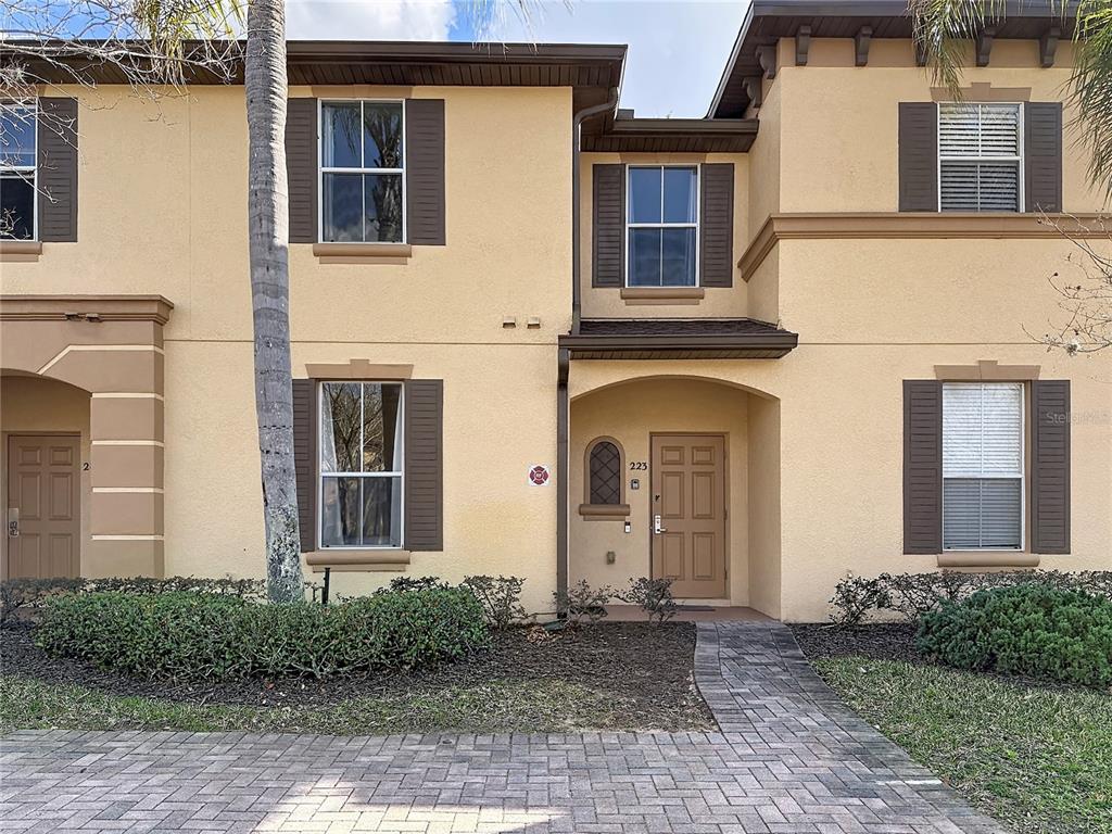 A two-story townhouse exterior features tan stucco walls, brown shutters, and a paved walkway — 223 NAPOLI DRIVE, DAVENPORT, FL