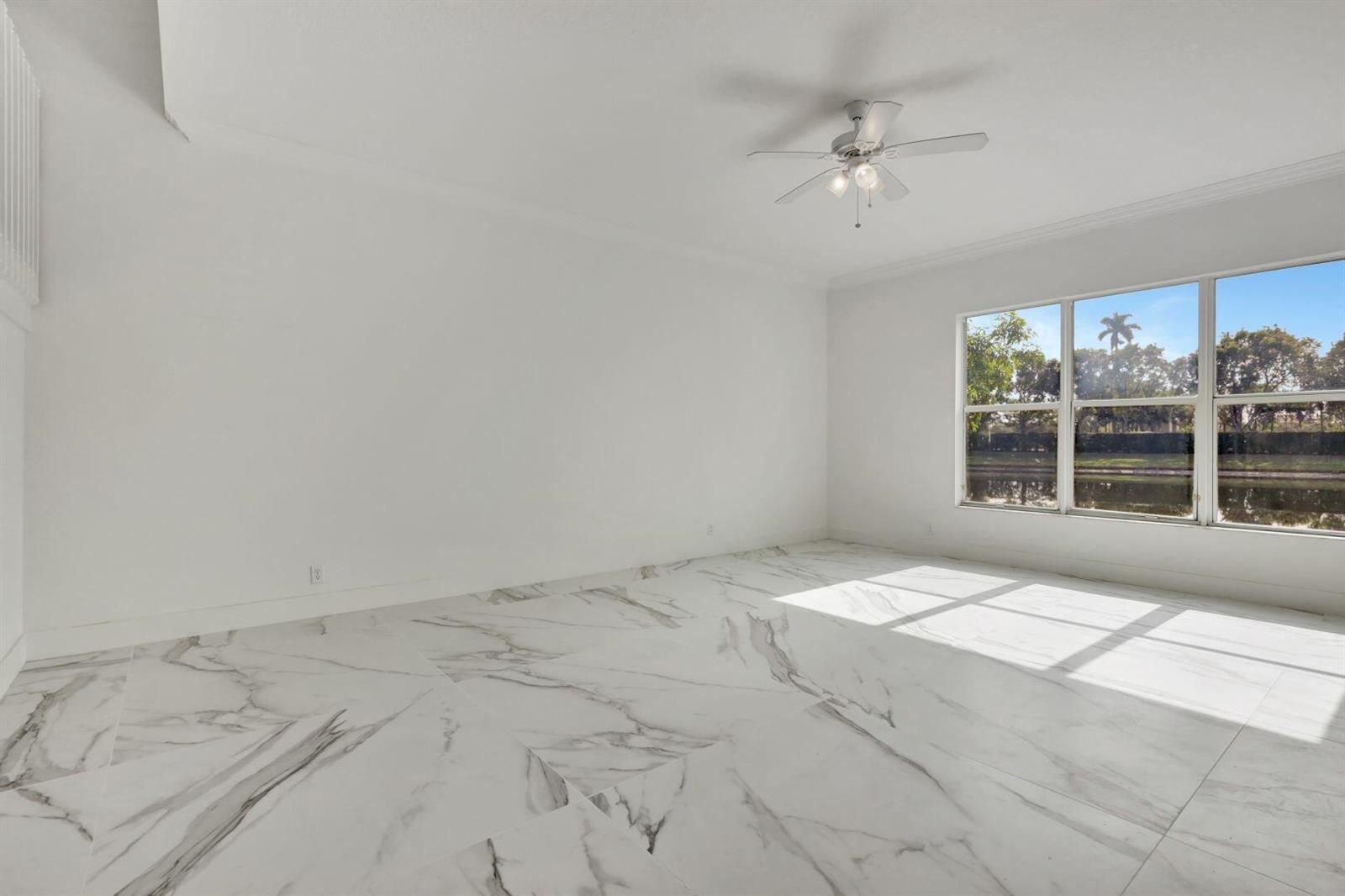 An empty room features white marble-look tile flooring, a ceiling fan, and large windows overlooking water — 9334 Madewood Court, Royal Palm Beach FL 33411, ROYAL PALM BEACH, FL