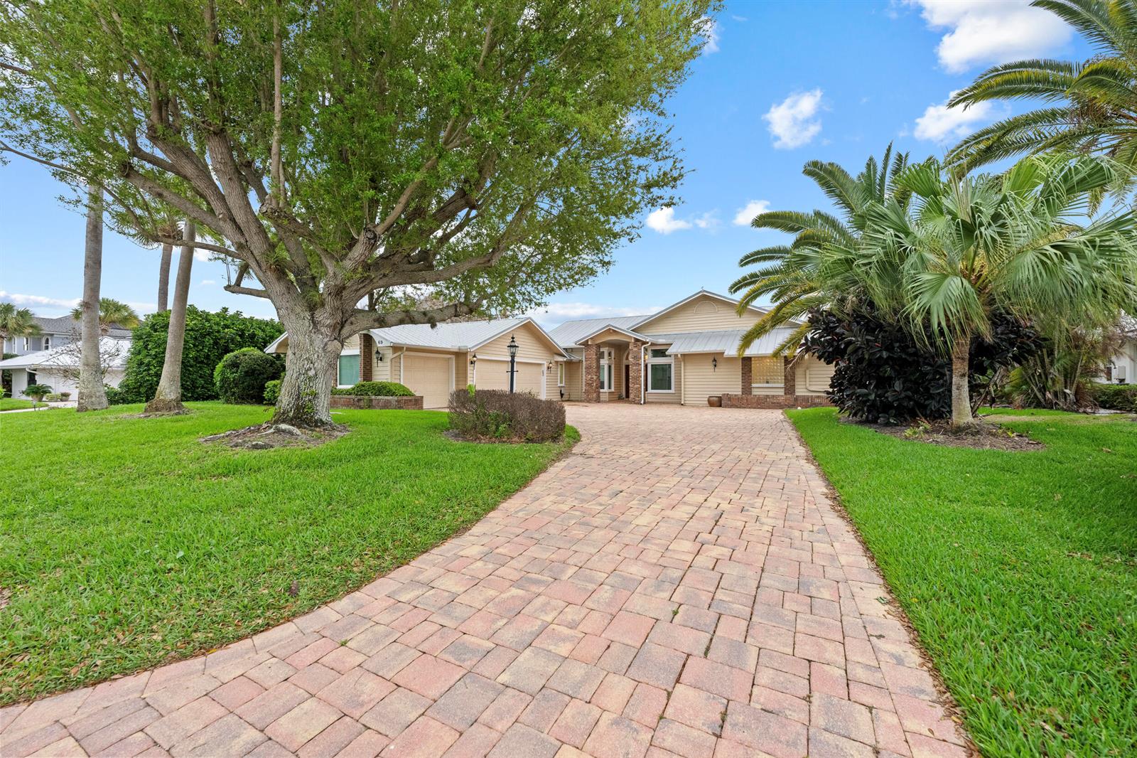 A beige house with a paver driveway, large tree, and palm trees under a blue sky — 69 Cache Cay Drive, Vero Beach FL 32963, VERO BEACH, FL
