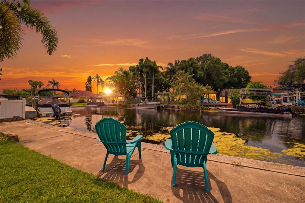 Two teal Adirondack chairs sit on a concrete patio overlooking a canal at sunset — 51 CYPRESS DRIVE, PALM HARBOR, FL