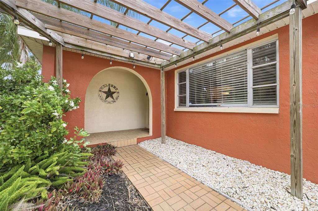 A terracotta-colored house exterior features a wooden pergola, a brick walkway, and a welcome sign — 51 CYPRESS DRIVE, PALM HARBOR, FL
