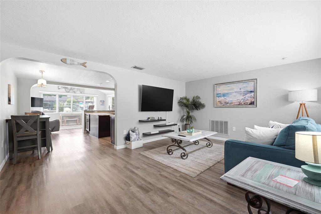 A living room with wood-look flooring, a wall-mounted television, and an open archway to a kitchen — 51 CYPRESS DRIVE, PALM HARBOR, FL