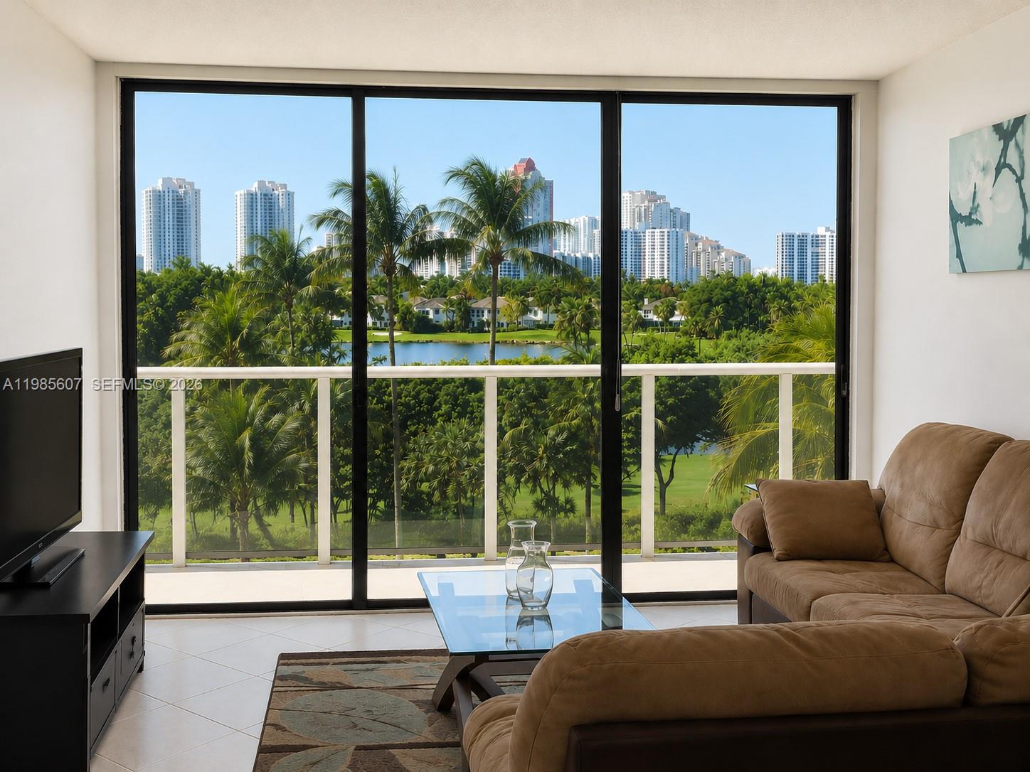 A living room features a brown sofa, glass coffee table, and sliding doors overlooking a lake — 3675 N Country Club Dr # 508, Aventura FL 33180, AVENTURA, FL