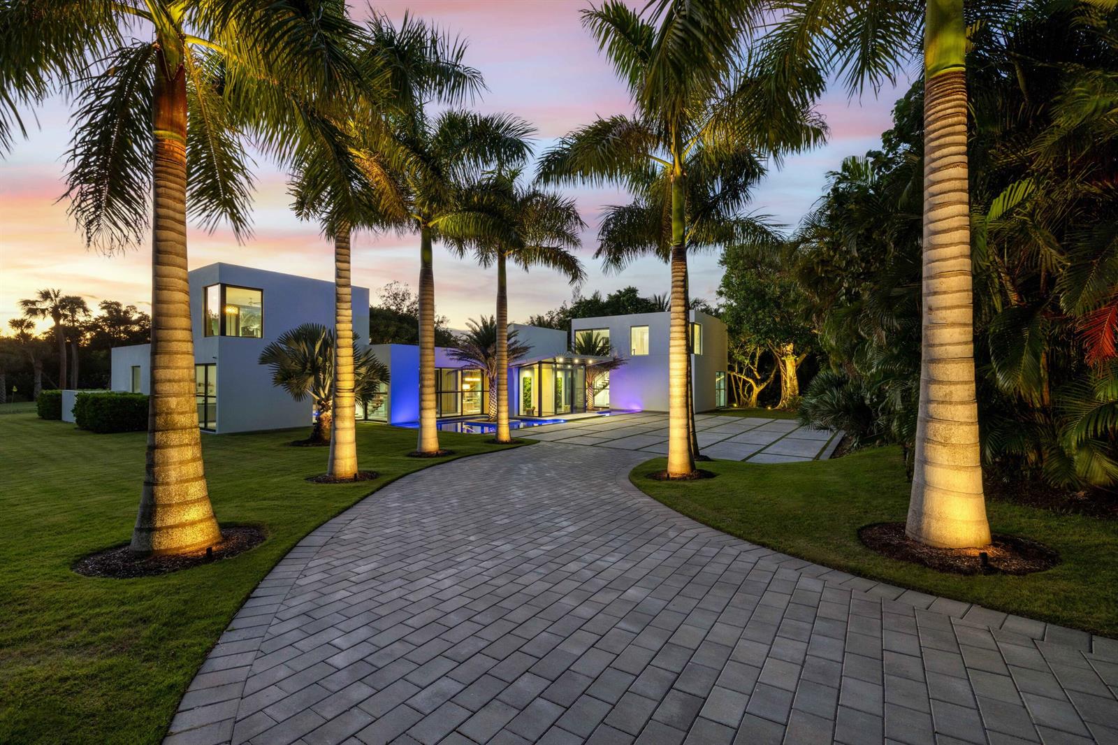 A modern white house with a paved driveway and illuminated palm trees at dusk — 12 N Beach Road, Jupiter Island FL 33455, JUPITER ISLAND, FL