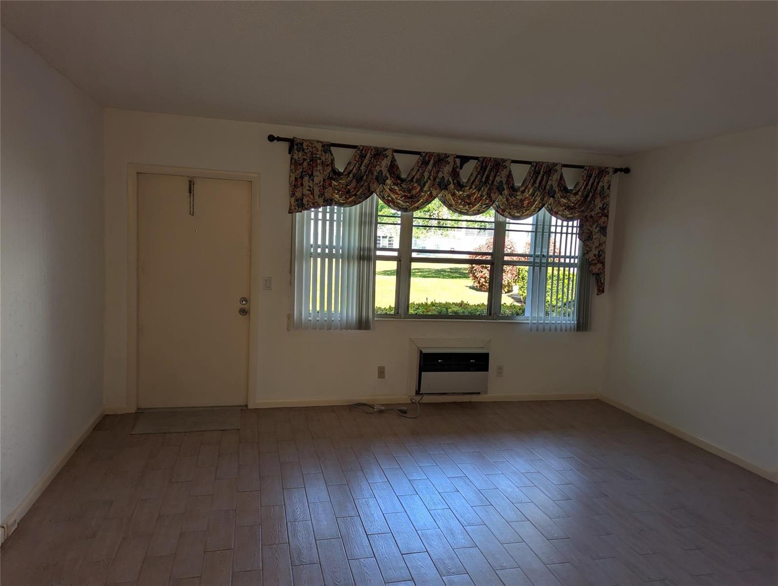 An empty living room features light wood-look tile flooring, a large window with floral valances, and an air conditioner — 190 Oakridge M, Deerfield Beach FL 33442, DEERFIELD BEACH, FL