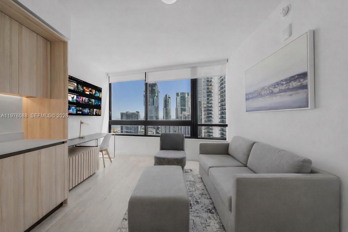 A modern living room features a gray sofa, a desk, and floor-to-ceiling windows with city views — 225 N Miami # 3009, Miami FL 33128, MIAMI, FL