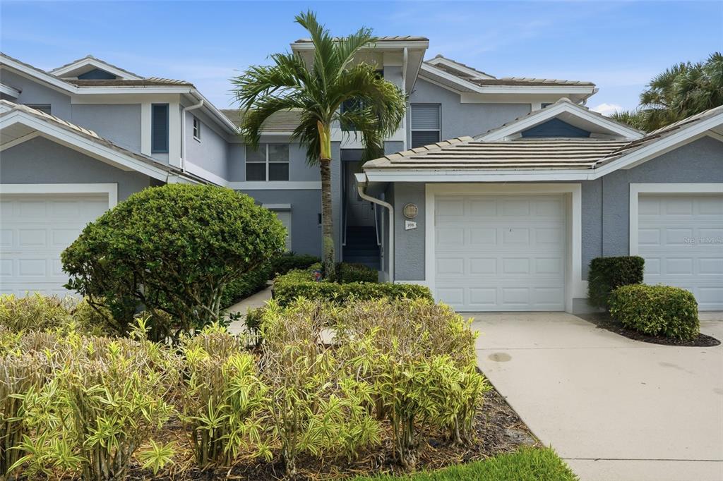A two-story residential building features a palm tree, manicured landscaping, and two white garage doors — 710 TARPON COVE DRIVE UNIT 203, NAPLES, FL