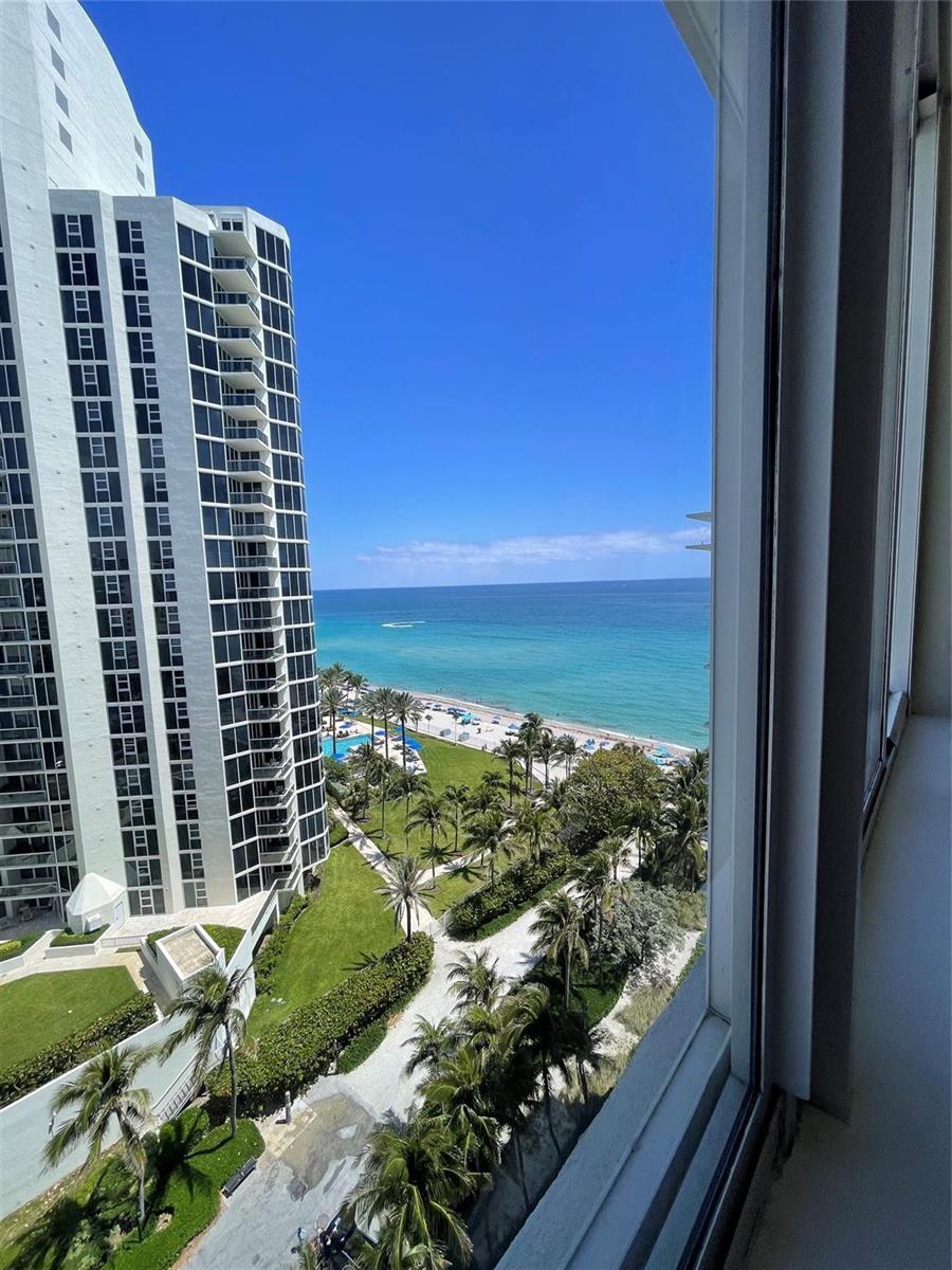 A high-angle view from a window overlooking a beach, palm trees, and a tall building — 19201 Collins Avenue, Sunny Isles Beach FL 33160, SUNNY ISLES BEACH, FL