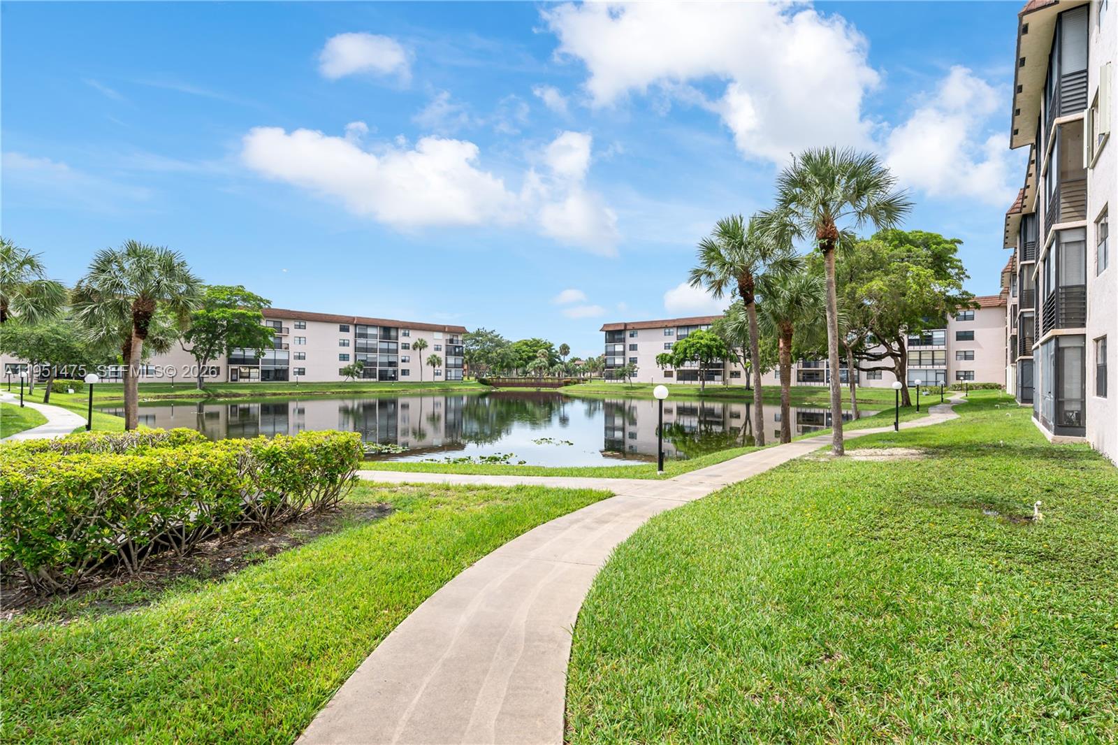 A concrete walkway winds through a grassy courtyard toward a pond surrounded by apartment buildings — 4960 E Sabal Palm Blvd # 301, Tamarac FL 33319, TAMARAC, FL