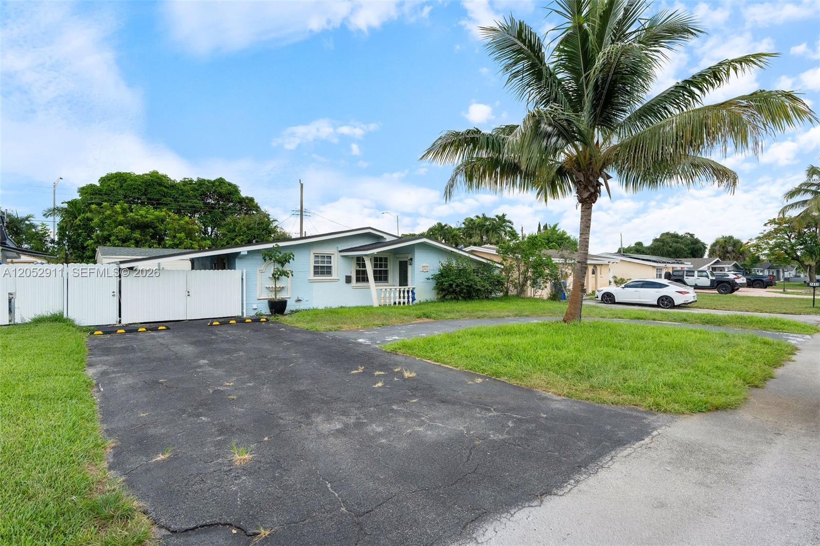 A light blue single-story house features a paved driveway, a white fence, and a palm tree — 7501 Granada Blvd, Miramar FL 33023, MIRAMAR, FL