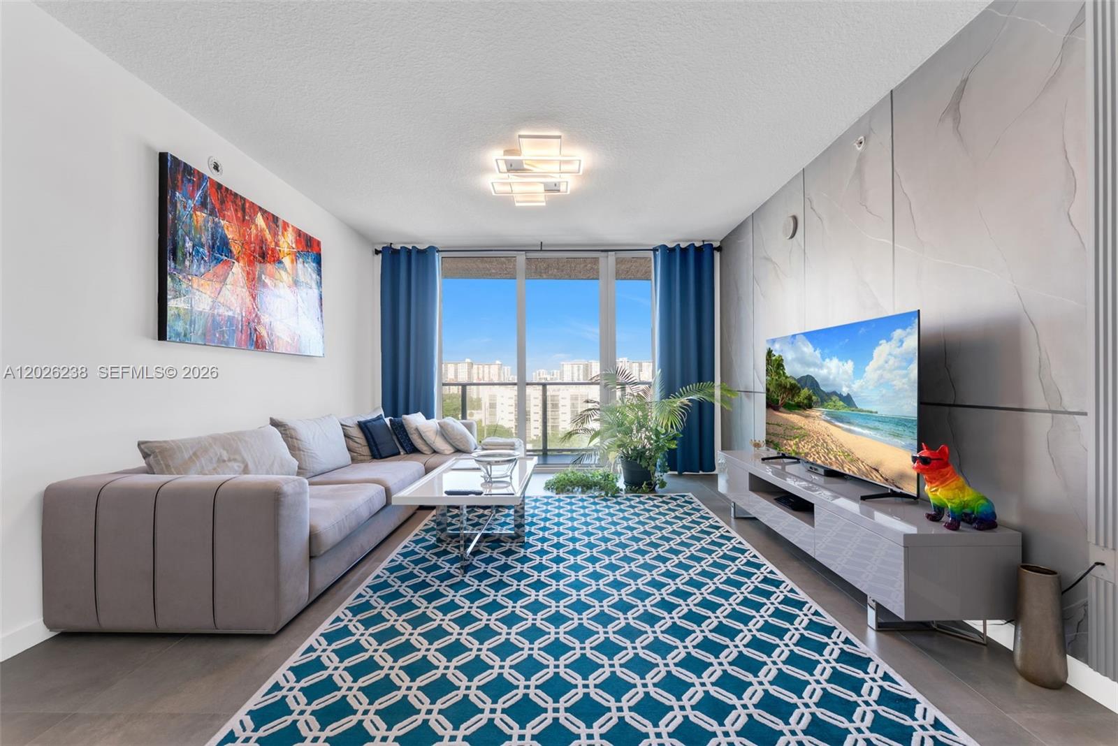 A modern living room features a gray sectional sofa, patterned rug, and a wall-mounted television — 300 Sunny Isles Blvd # 905, Sunny Isles Beach FL 33160, SUNNY ISLES BEACH, FL
