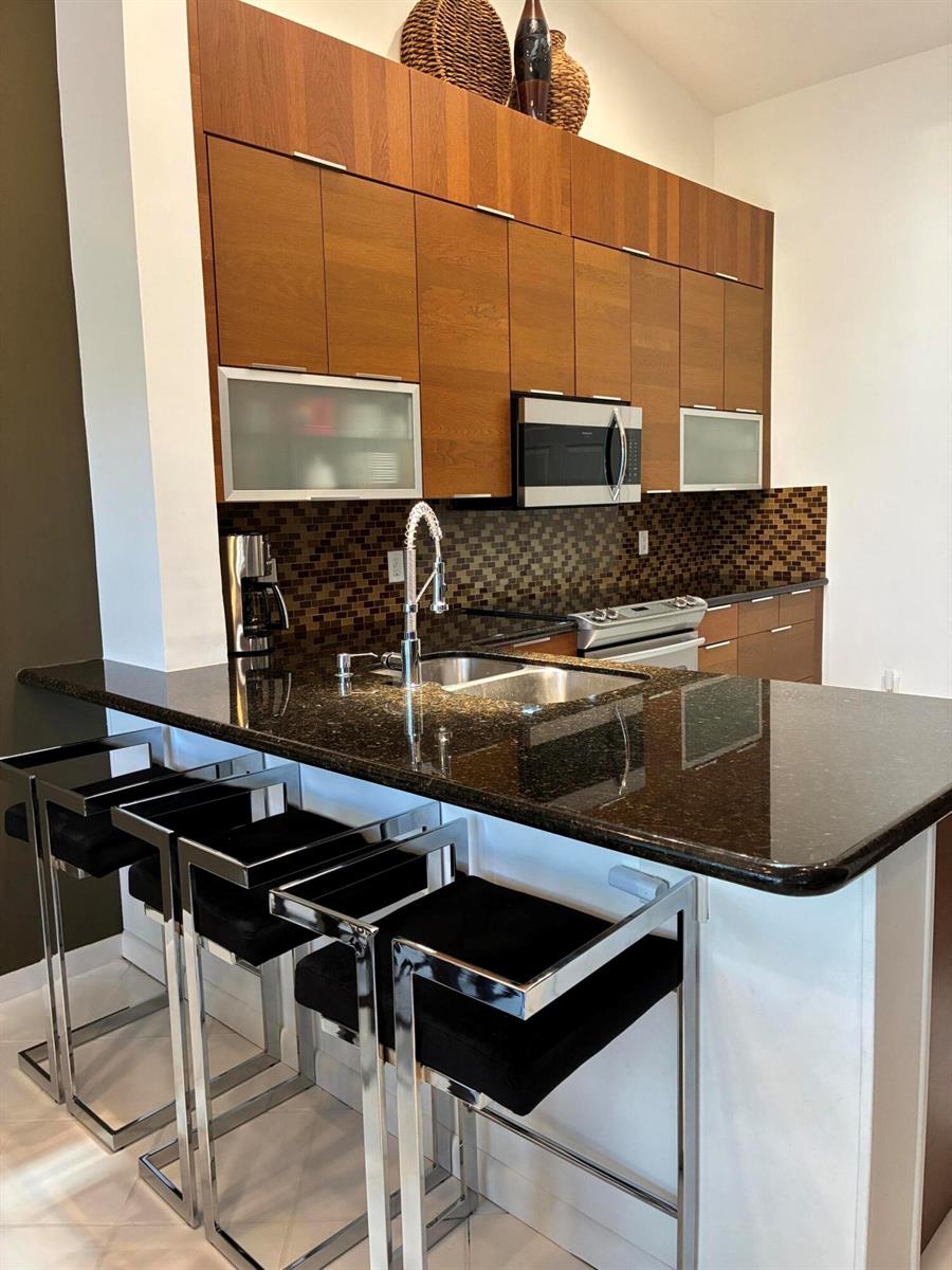 A modern kitchen features dark wood cabinetry, granite countertops, and three black bar stools — 17223 Boca Club Boulevard # 6, Boca Raton FL 33487, BOCA RATON, FL