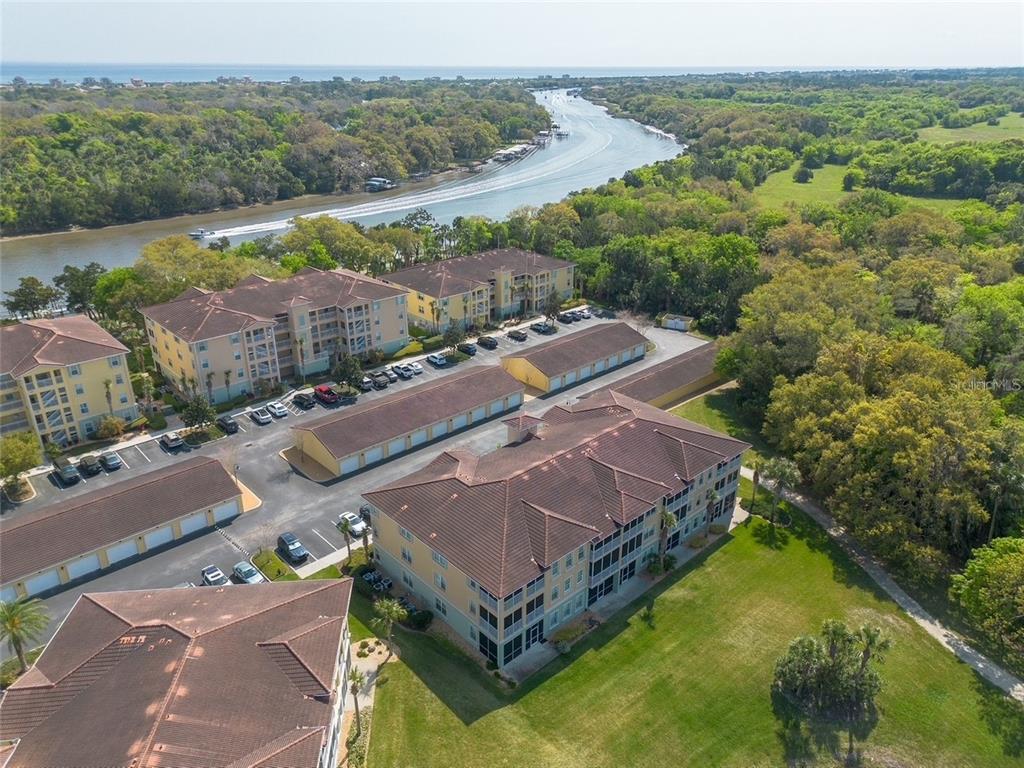 An aerial view shows multi-story apartment buildings with parking garages near a river and forest — 1000 CANOPY WALK LANE UNIT 1013, PALM COAST, FL