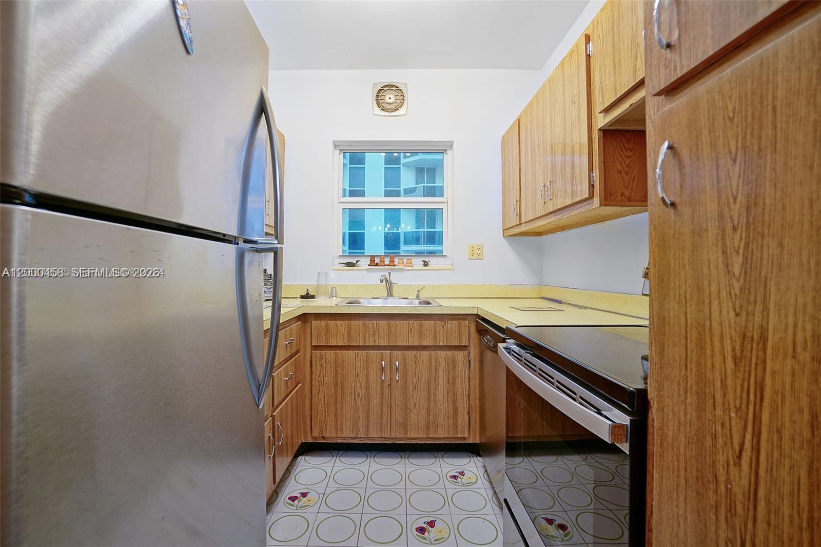 A galley kitchen features wood cabinets, a stainless steel refrigerator, and patterned tile flooring — 4747 Collins Ave # 1108, Miami Beach FL 33140, MIAMI BEACH, FL