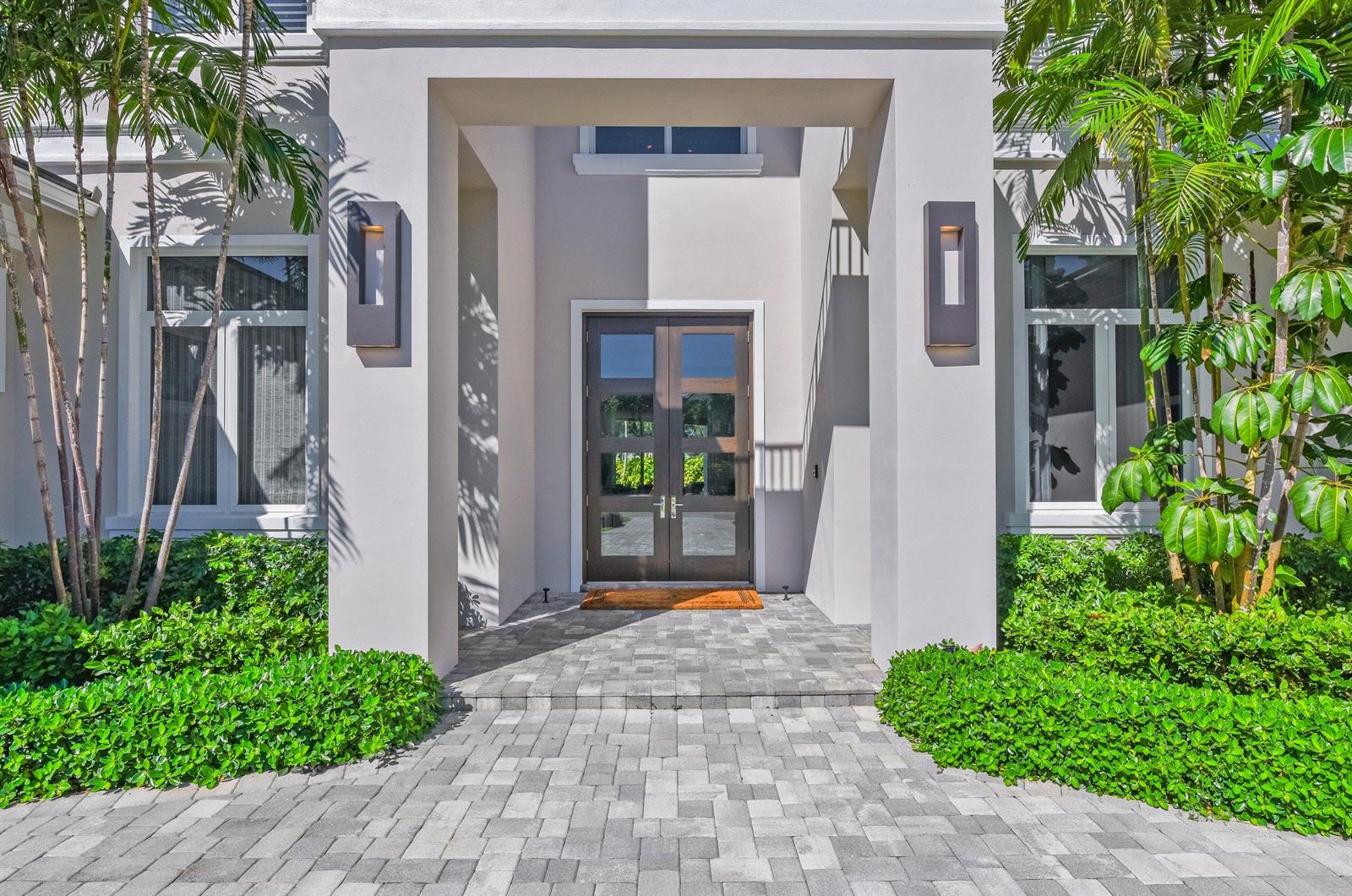 A modern home entrance features a double wood door, stone pavers, and lush green landscaping — 17661 Scarsdale Way, Boca Raton FL 33496, BOCA RATON, FL