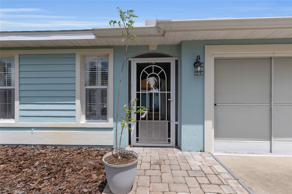 A light blue house exterior features a white storm door, a potted plant, and pavers — 7221 SW 115TH PLACE, OCALA, FL