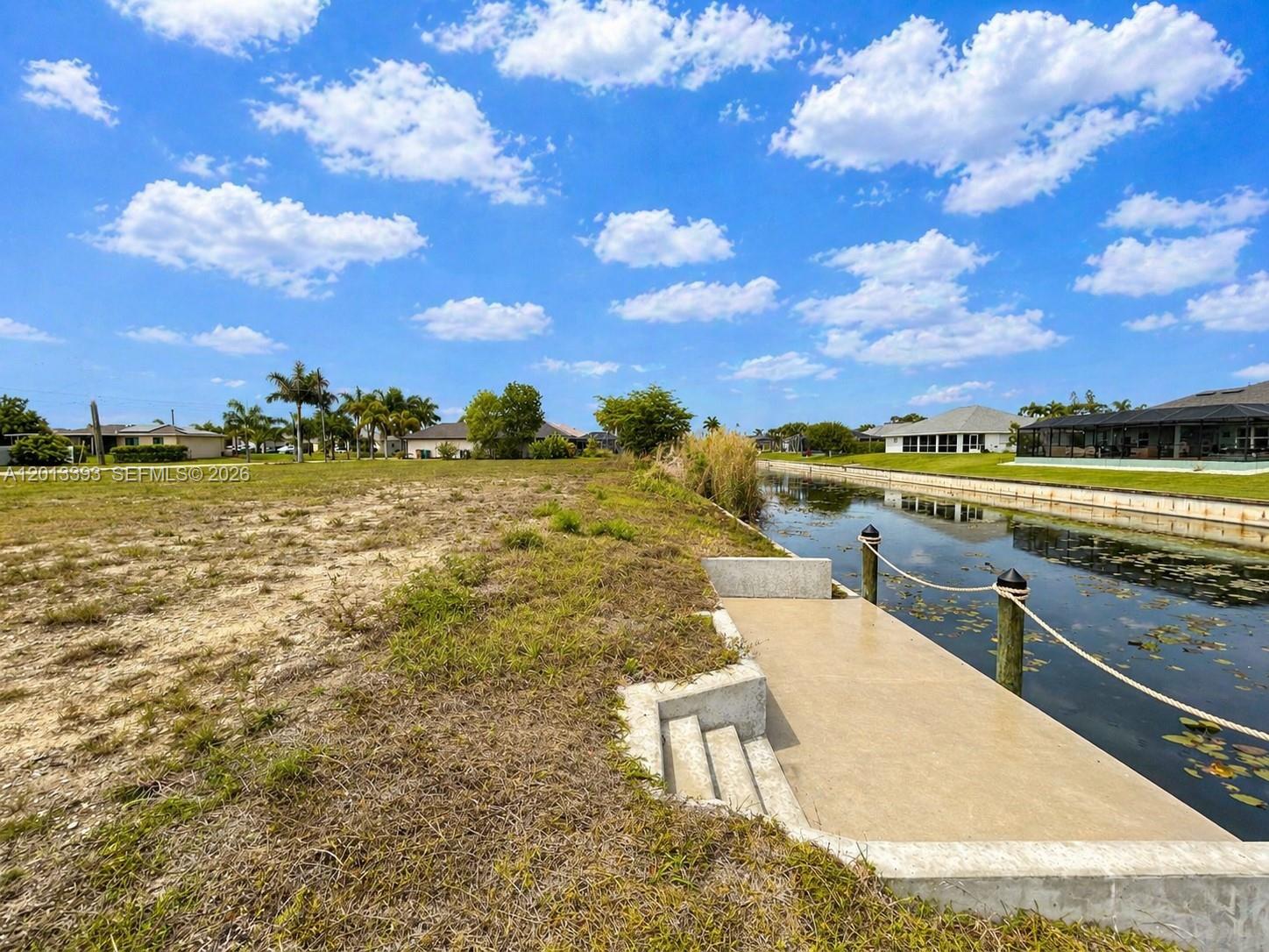 A vacant grassy lot features a concrete dock extending into a canal under a blue sky — 1014 SW 18th St, Cape Coral FL 33991, CAPE CORAL, FL