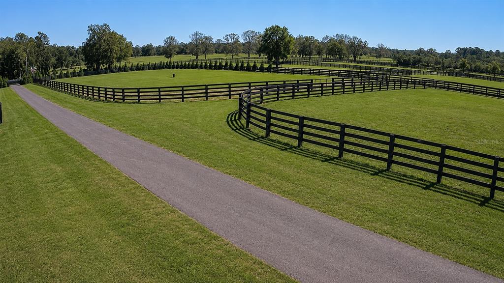 An aerial view shows a paved driveway winding through green pastures enclosed by black fencing — 8020 NW 115TH AVENUE, OCALA, FL