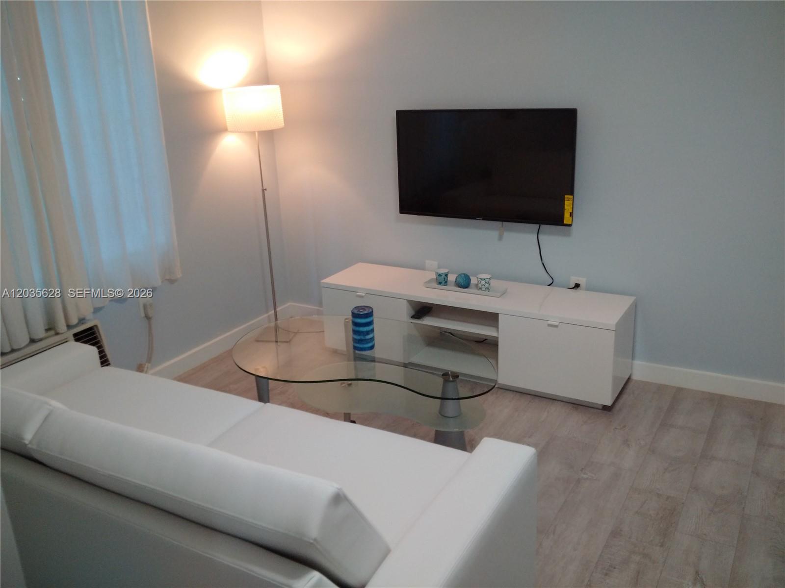 A living room features a white sofa, glass coffee table, media console, and wall-mounted television — 1525 Meridian Ave # 104, Miami Beach FL 33139, MIAMI BEACH, FL