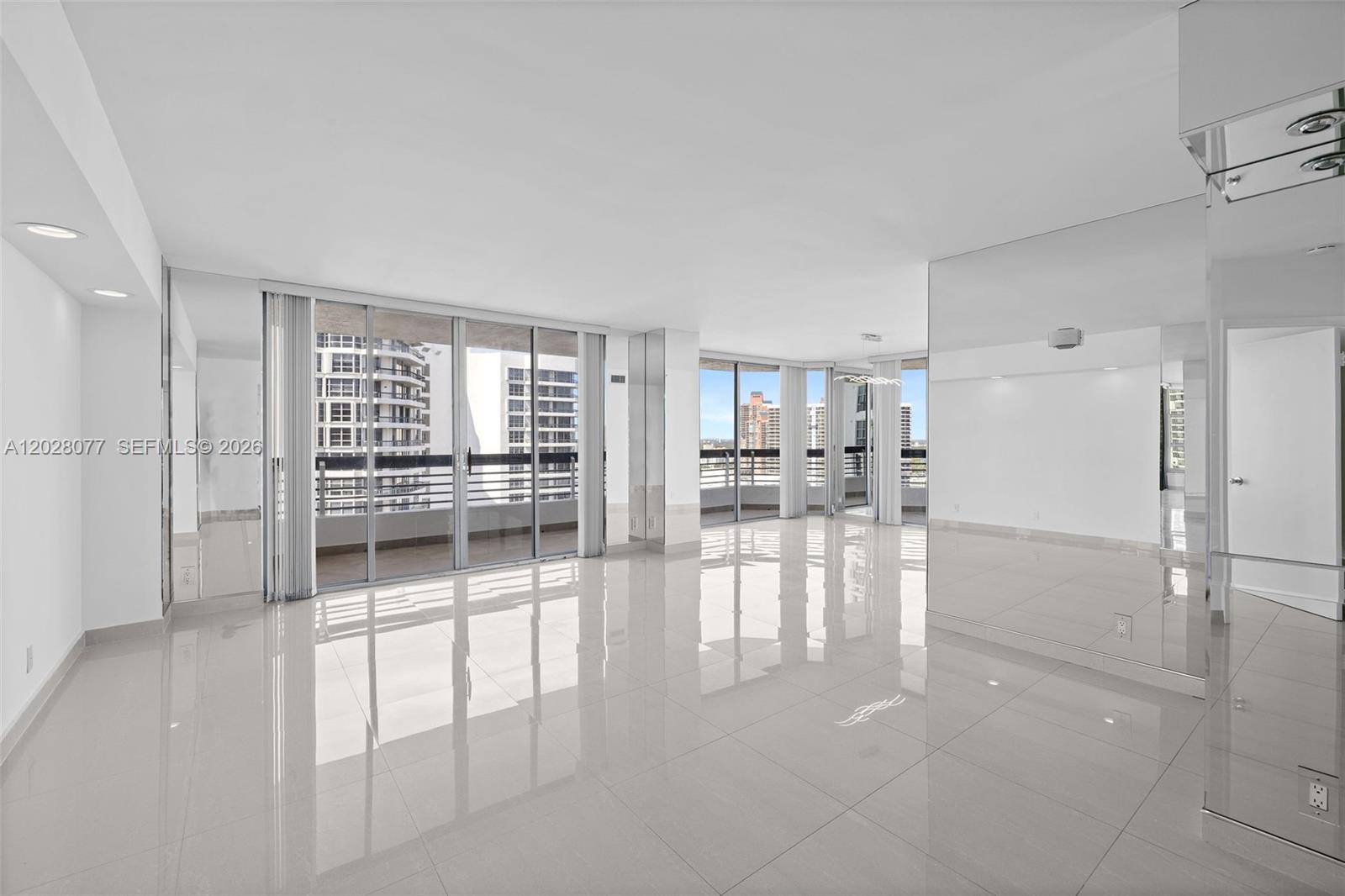 A bright, empty living room features white tile floors, floor-to-ceiling windows, and mirrored walls — 19195 NE 36th Ct # 2705, Aventura FL 33180, AVENTURA, FL