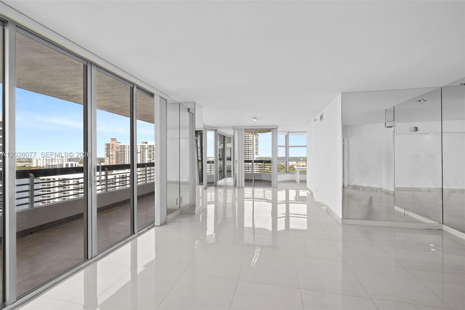 An empty room with white tile floors, mirrored walls, and floor-to-ceiling windows overlooking a balcony — 19195 NE 36th Ct # 2705, Aventura FL 33180, AVENTURA, FL