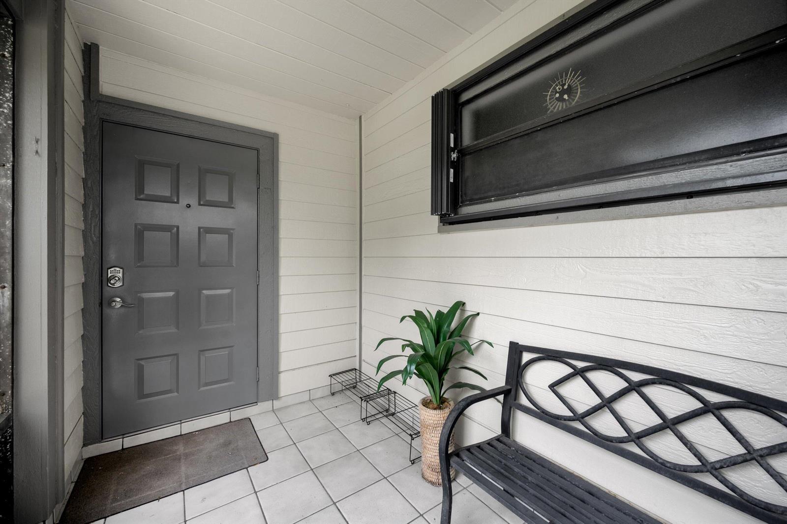 A covered entryway features a gray door, a metal bench, a potted plant, and tile flooring — 2482 Bedford Mews Drive, Wellington FL 33414, WELLINGTON, FL