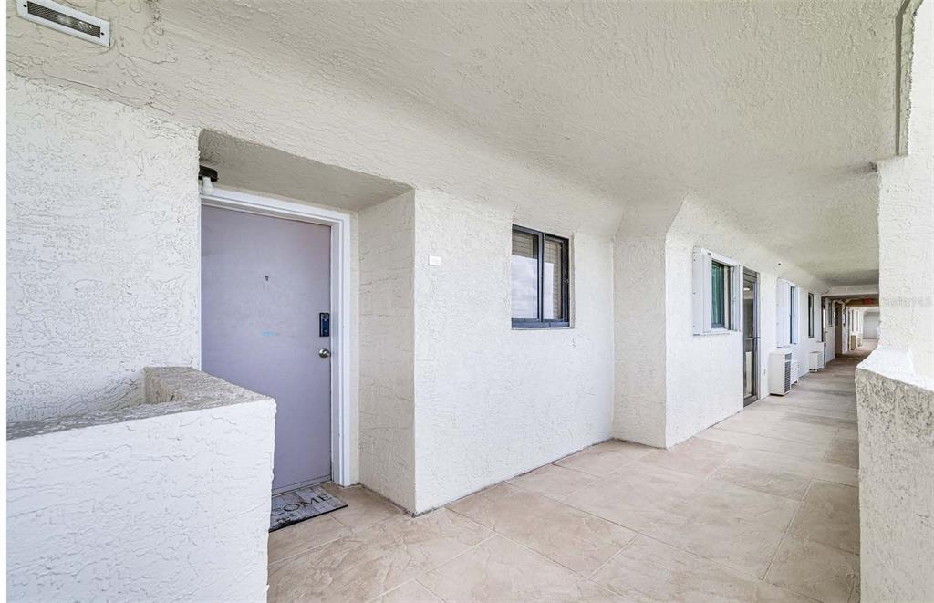 An outdoor apartment hallway features a light-colored door, tiled flooring, and white stucco walls — 5915 SEA RANCH DRIVE UNIT 310, HUDSON, FL