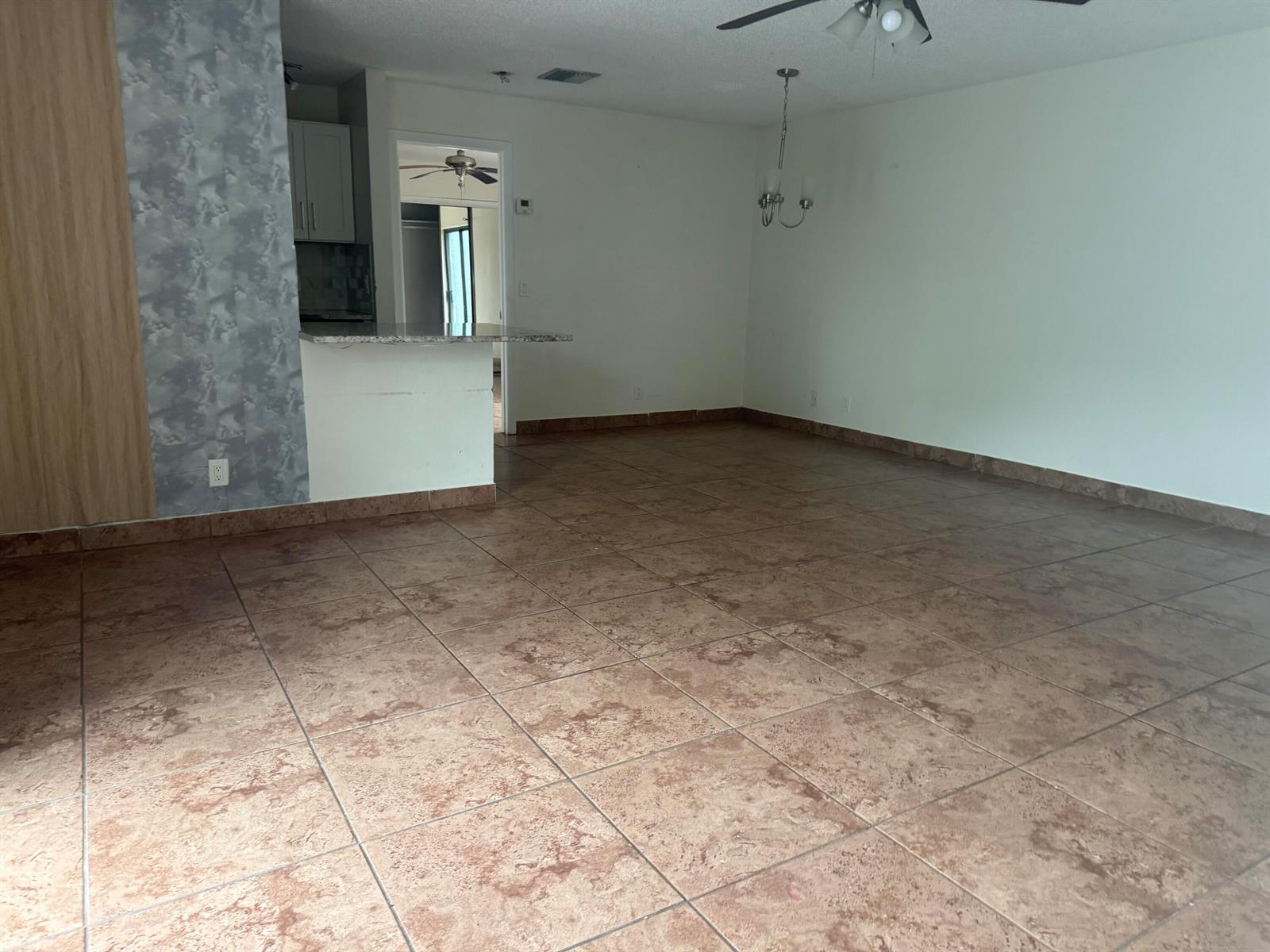 An empty room features large brown floor tiles, a kitchen pass-through, and a ceiling fan — 21 Crossings Circle # C, Boynton Beach FL 33435, BOYNTON BEACH, FL