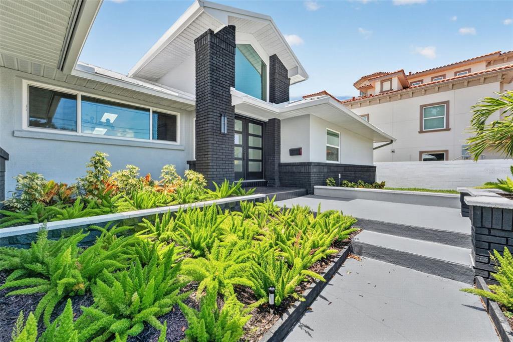 A modern house exterior features a dark brick chimney, a glass front door, and landscaped gardens — 2936 68TH AVENUE S, ST PETERSBURG, FL