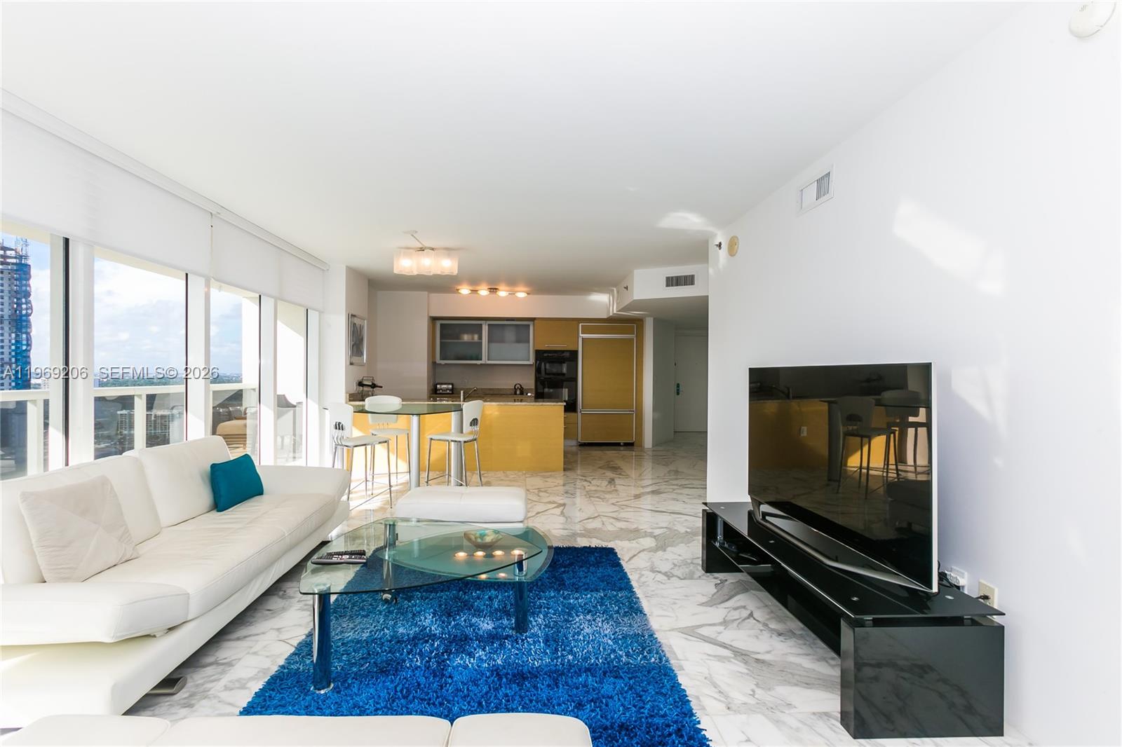 A modern living room features white leather furniture, a blue rug, and an open kitchen — 1830 S OCEAN DR # 2906, Hallandale Beach FL 33009, HALLANDALE BEACH, FL