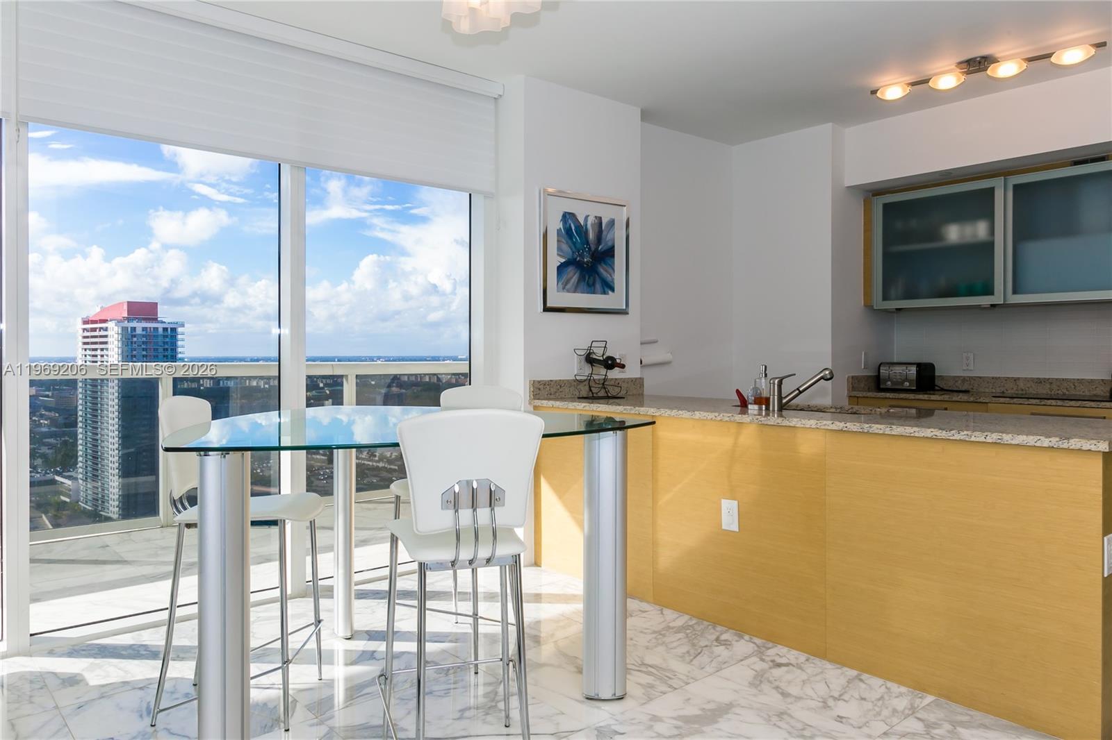 A kitchen with light wood cabinetry and a glass-topped dining table overlooks a city skyline — 1830 S OCEAN DR # 2906, Hallandale Beach FL 33009, HALLANDALE BEACH, FL