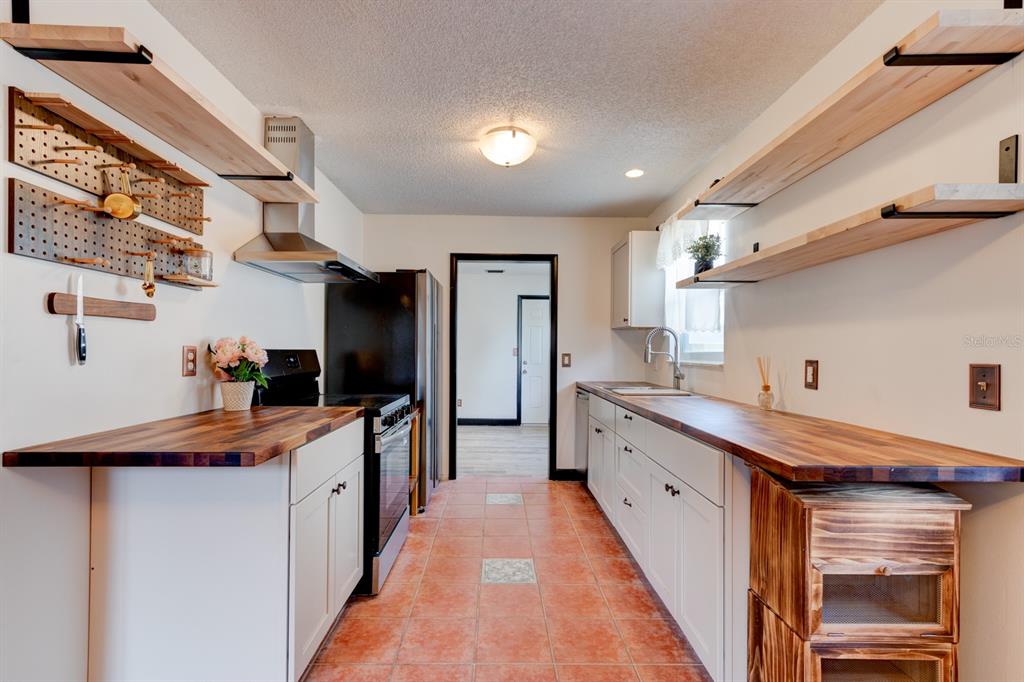 A galley kitchen features white cabinets, butcher block countertops, open shelving, and terracotta tile flooring — 119 BIGELOW DRIVE, EDGEWATER, FL