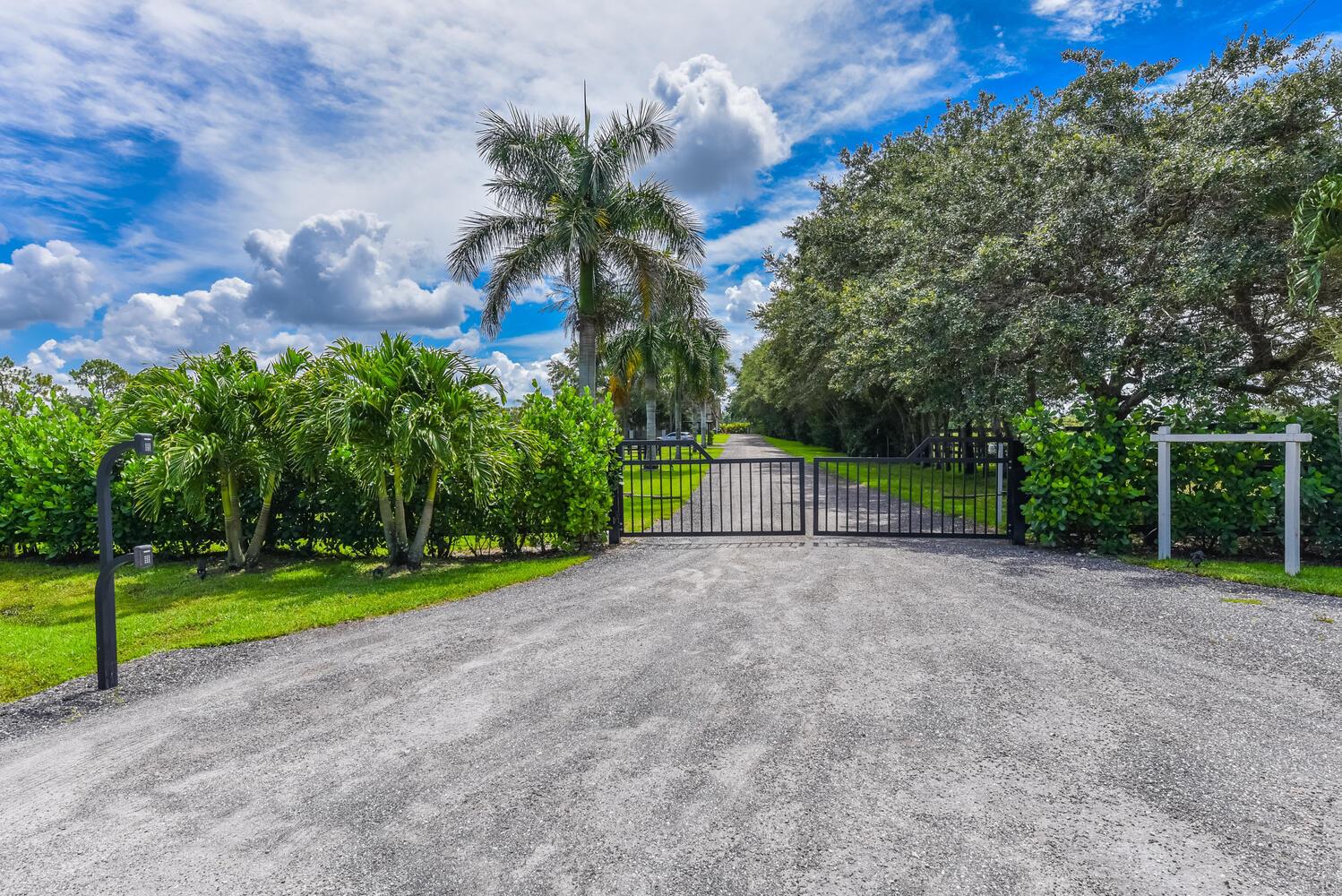 A gravel driveway leads to a black metal gate flanked by palm trees and landscaping — 2780 Skipiks Way, Wellington FL 33470, WELLINGTON, FL