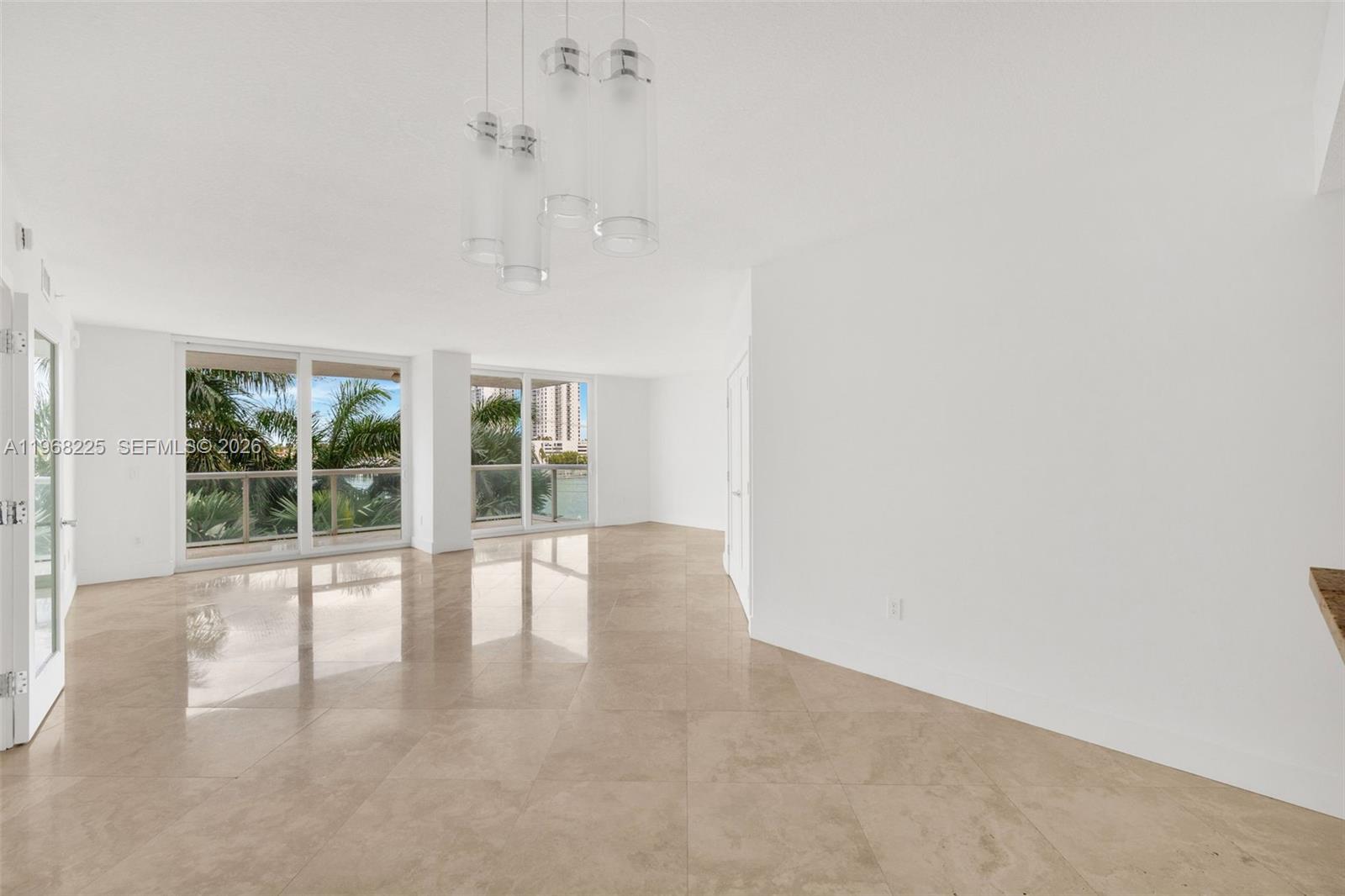 A bright, empty living room features large windows, beige tile flooring, and a modern chandelier — 10 Venetian Way # 403, Miami Beach FL 33139, MIAMI BEACH, FL