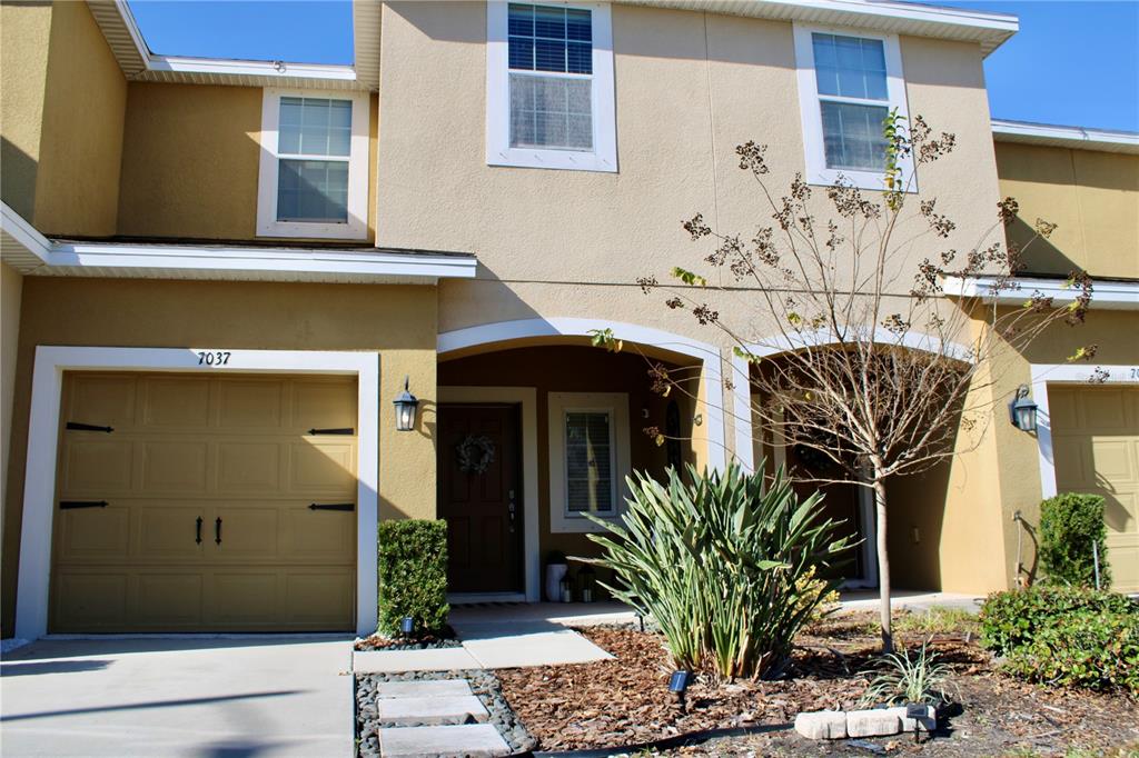 The exterior of a two-story tan townhouse features a garage, a covered entryway, and landscaping — 7037 WOODCHASE GLEN DRIVE, RIVERVIEW, FL