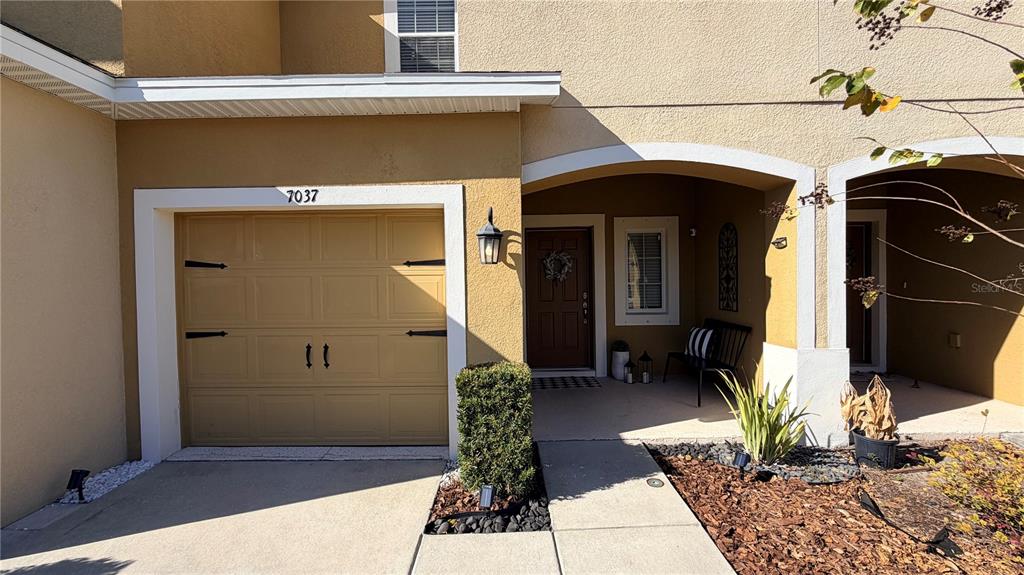 A tan stucco townhouse exterior features a single-car garage, a covered entryway, and landscaping — 7037 WOODCHASE GLEN DRIVE, RIVERVIEW, FL