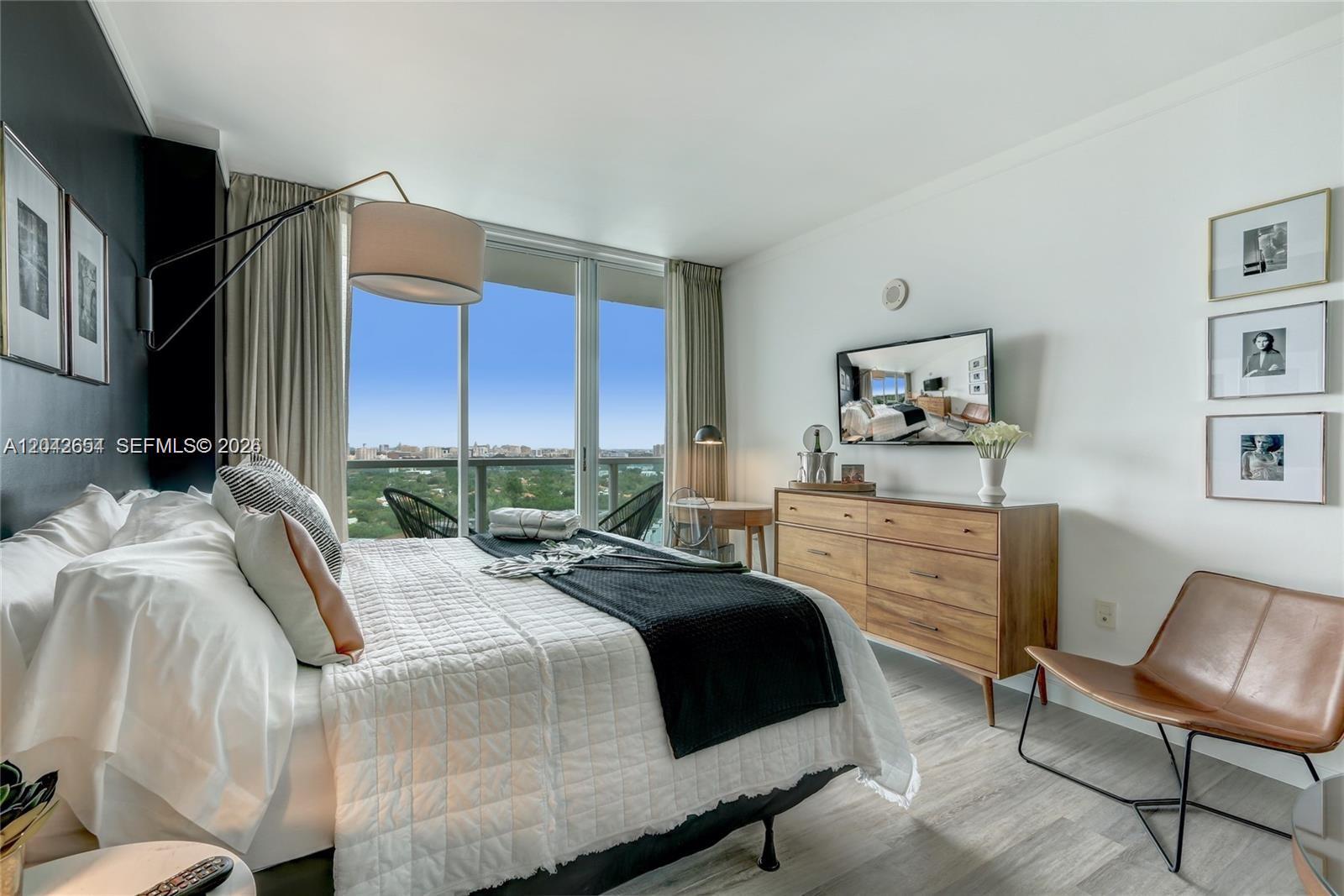 A bedroom features a bed, wooden dresser, wall-mounted television, and floor-to-ceiling windows with balcony views — 2889 Mcfarlane Rd # 1811, Miami FL 33133, MIAMI, FL