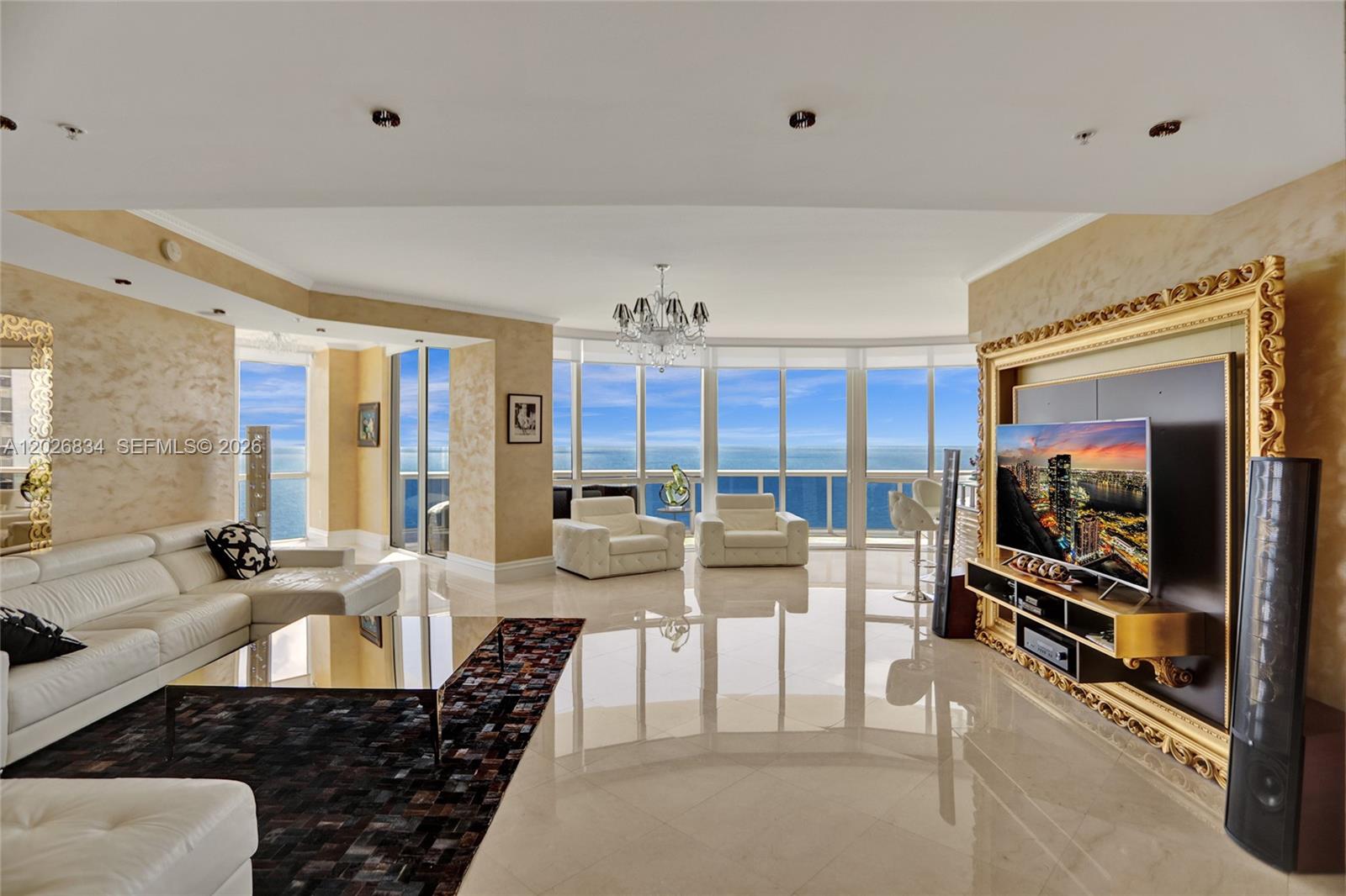 A bright living room features white leather furniture, a gold-framed television, and floor-to-ceiling ocean views — 18201 Collins Ave # 3309, Sunny Isles Beach FL 33160, SUNNY ISLES BEACH, FL
