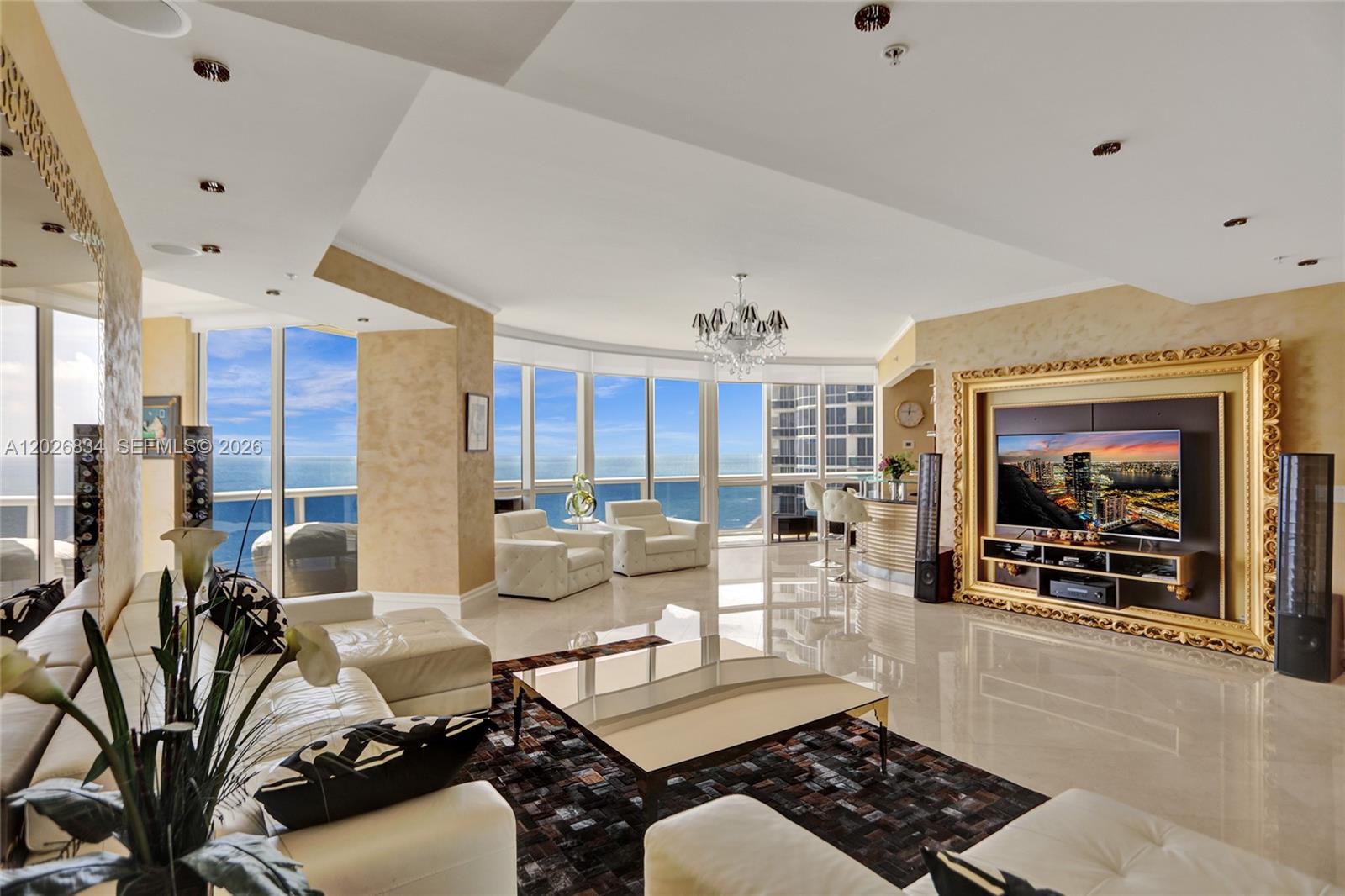 A modern living room features white leather furniture, a gold-framed television, and panoramic ocean views — 18201 Collins Ave # 3309, Sunny Isles Beach FL 33160, SUNNY ISLES BEACH, FL