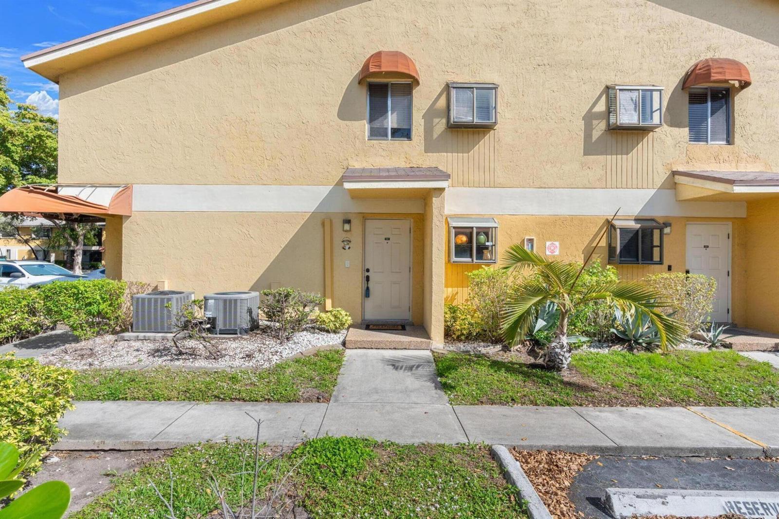 An exterior view of a two-story tan stucco townhouse with a concrete walkway and landscaping — 3053 N Oakland Forest Drive # 102, Oakland Park FL 33309, OAKLAND PARK, FL