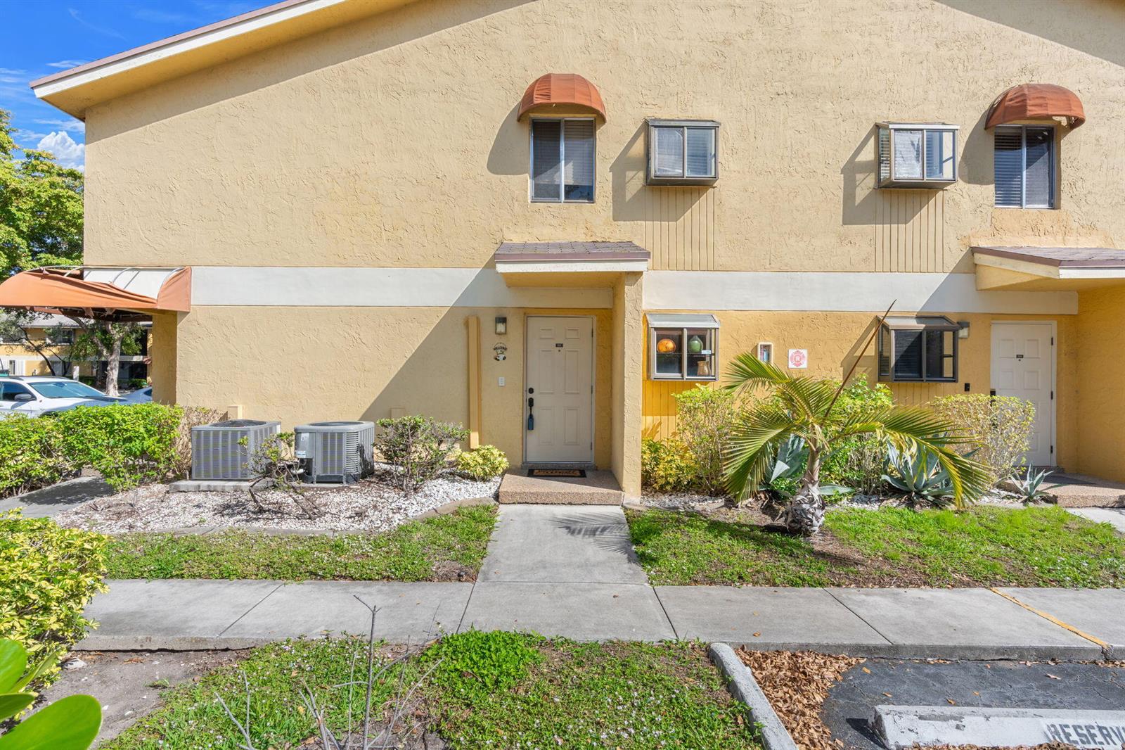The exterior of a two-story yellow stucco townhouse features a concrete walkway and landscaping — 3053 N Oakland Forest Drive # 102, Oakland Park FL 33309, OAKLAND PARK, FL