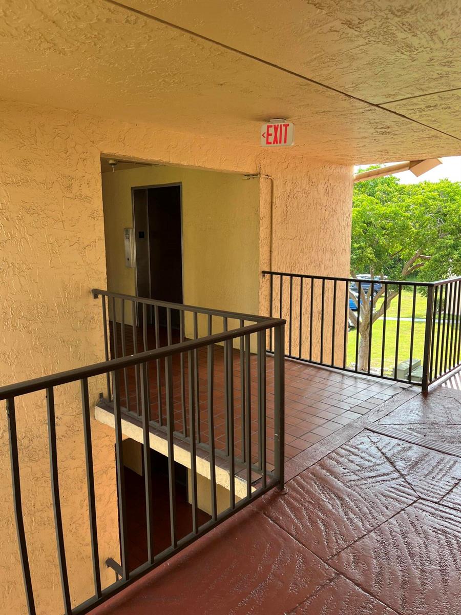 An outdoor apartment building walkway features a metal railing, tiled flooring, and an elevator entrance — 3396 Foxcroft Road # 309, Miramar FL 33025, MIRAMAR, FL