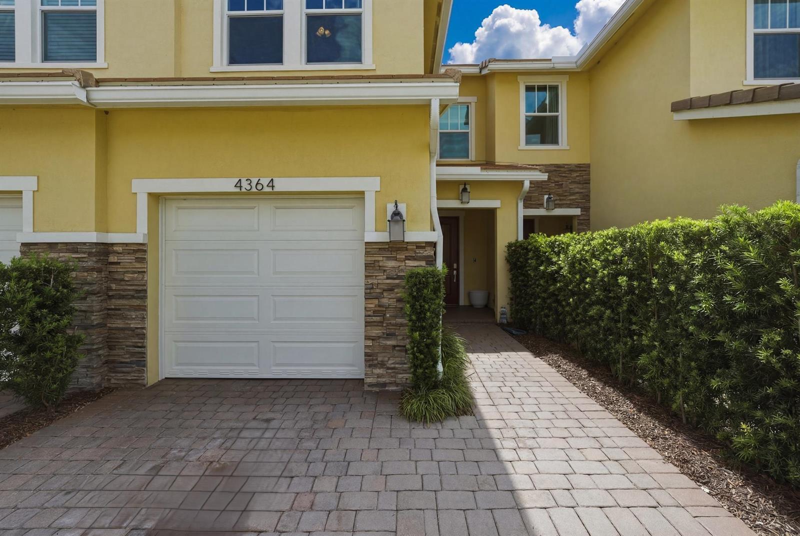 A yellow two-story townhouse features a white garage door, stone accents, and a paved walkway — 4364 Veleiros Avenue, Deerfield Beach FL 33064, DEERFIELD BEACH, FL