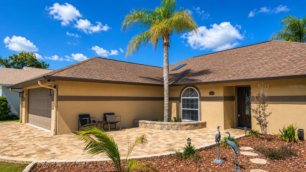 A tan house features a paved driveway, a palm tree, and two chairs on the patio — 6441 SILVER OAKS DRIVE, ZEPHYRHILLS, FL