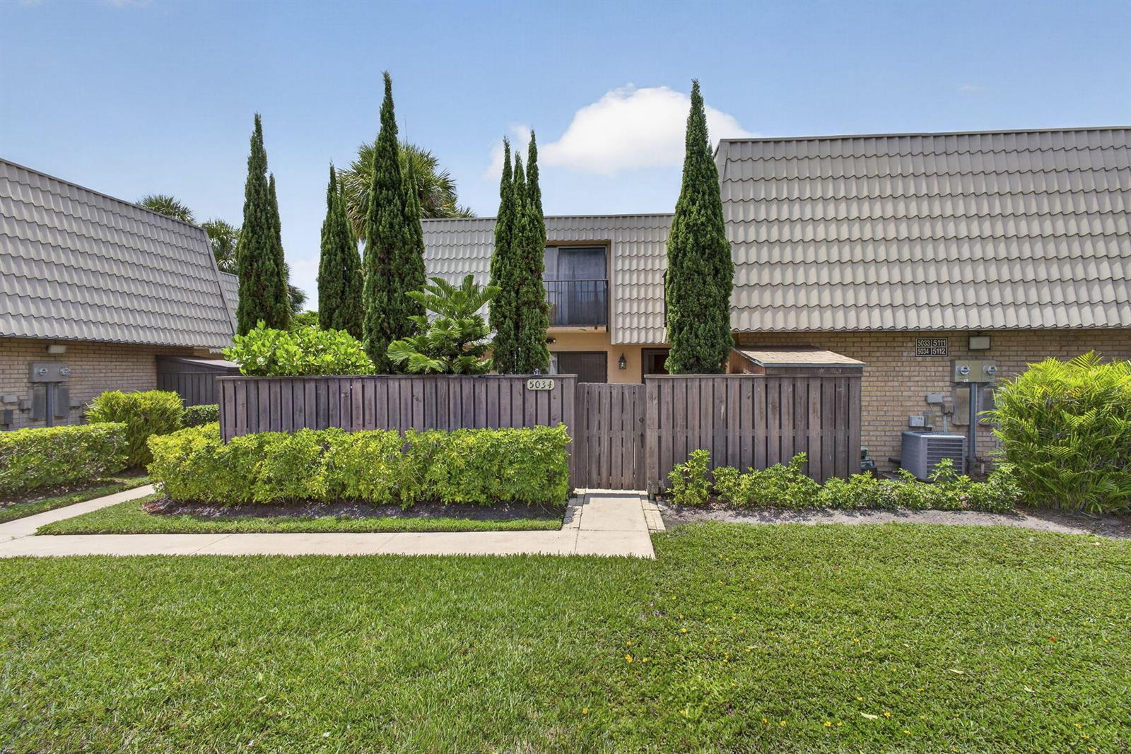 A two-story townhouse exterior features a wooden privacy fence, tall cypress trees, and landscaping — 5034 50th Way, West Palm Beach FL 33409, WEST PALM BEACH, FL