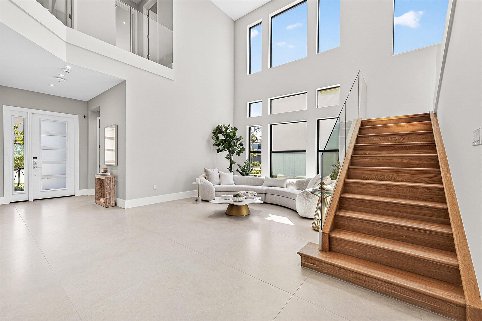 A bright, high-ceilinged foyer features a curved sofa, wooden staircase, and large windows — 9370 Sunflower Meadow Road, Boca Raton FL 33434, BOCA RATON, FL