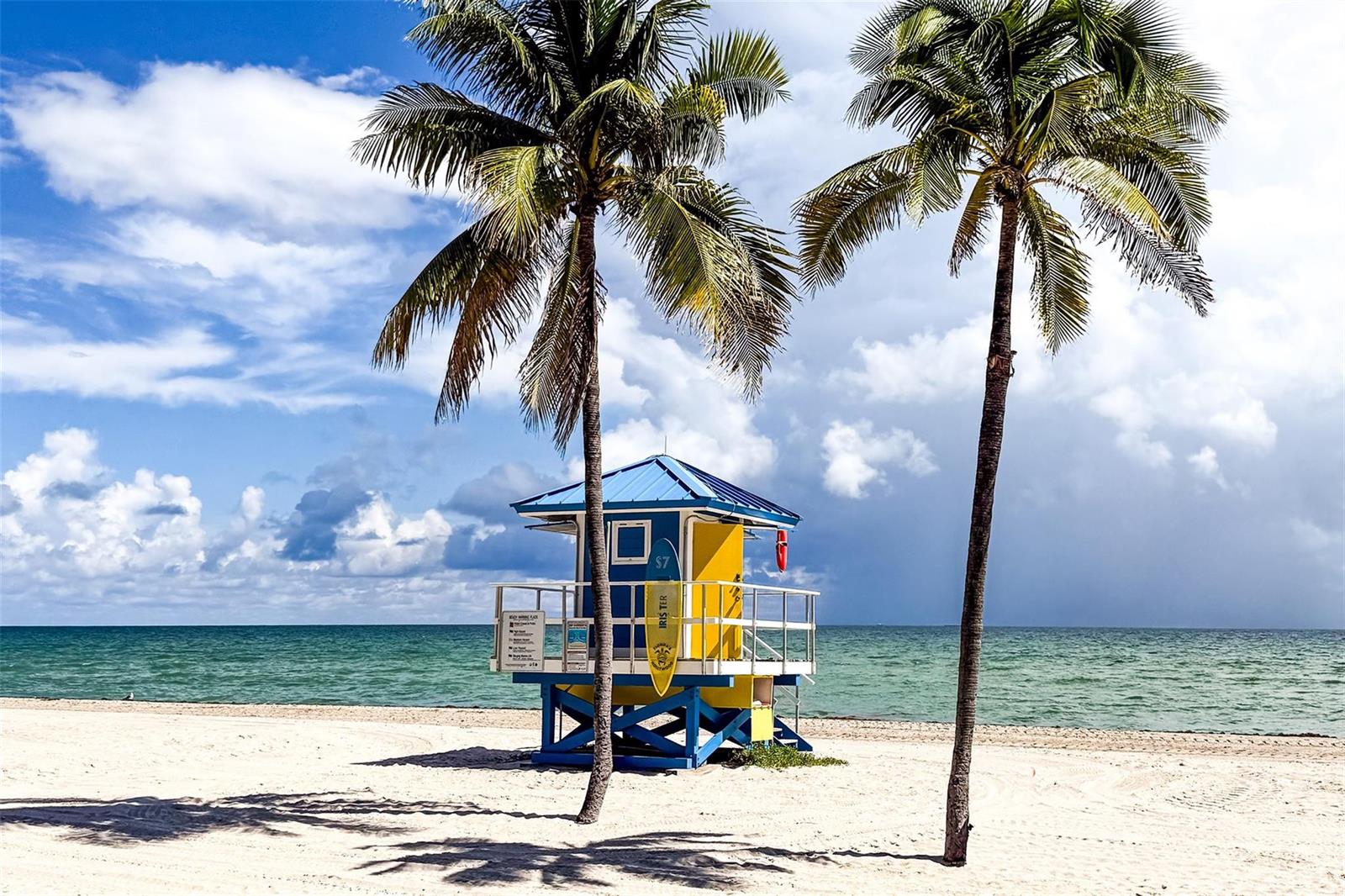 A yellow and blue lifeguard tower stands on a sandy beach between two palm trees — 2205 S Surf Road # 4b, Hollywood FL 33019, HOLLYWOOD, FL