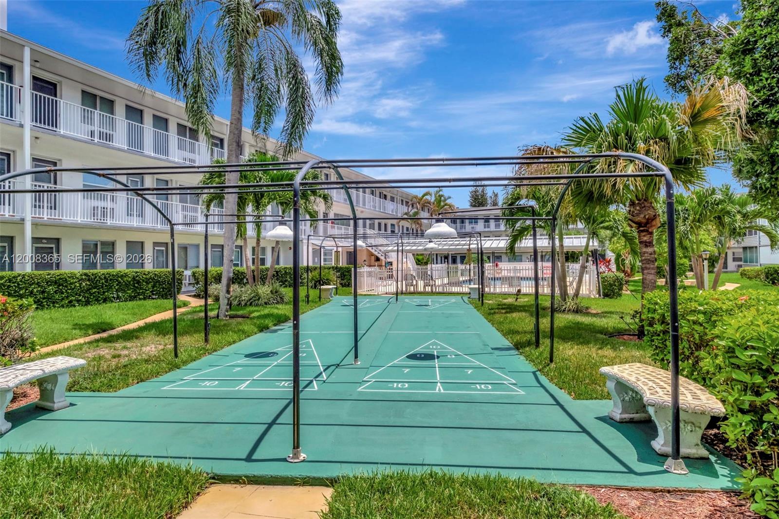 A green shuffleboard court sits outdoors between two white apartment buildings with palm trees — 15610 NE 6th Ave # 23C, Miami FL 33162, MIAMI, FL