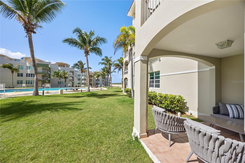 A covered patio with outdoor furniture overlooks a grassy courtyard with palm trees and buildings — Haudimar Beach Apartments 400 4466 ROAD UNIT H102, ISABELA, PR